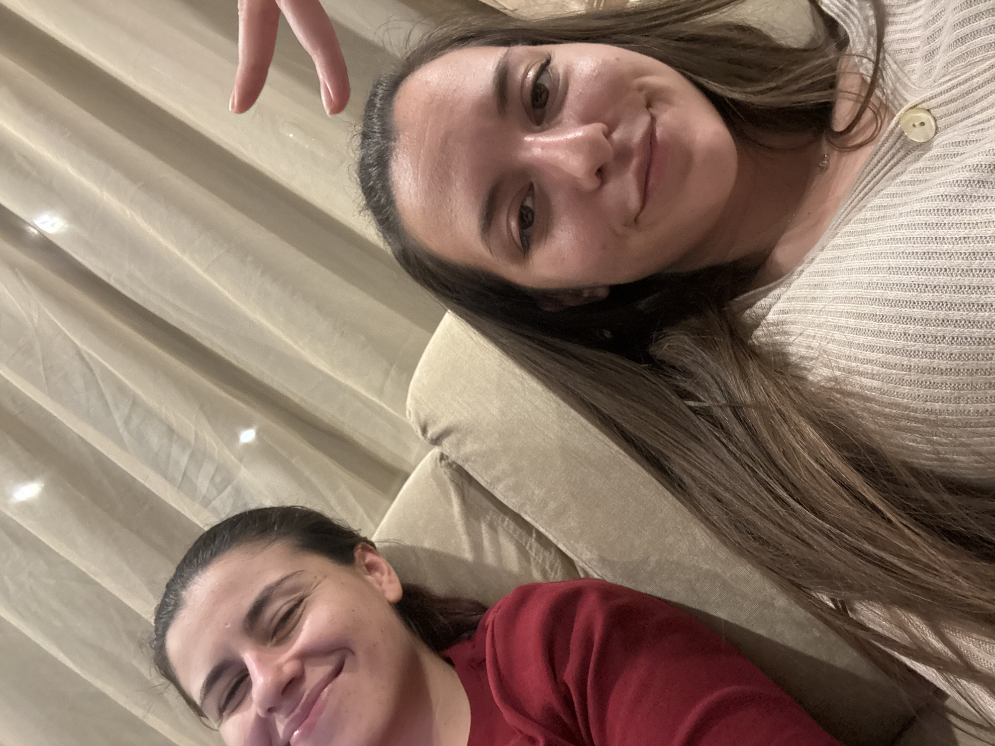 Two young adult females are depicted indoors, seated on a light beige fabric-upholstered couch. The female on the left is partially visible, facing slightly right, wearing a red long-sleeved garment, and smiling with her eyes mostly closed. The female on the right is centrally positioned, looking towards the camera with a subtle smile. She has long brown hair and is wearing a light beige ribbed long-sleeved top with a single button visible near the neckline; a delicate silver necklace is also visible. Her right hand, with the index and middle fingers extended in a gesture, is partially visible in the upper right. The background consists of light beige, vertically folded fabric drapes or curtains, featuring several bright reflective spots. The specific location within Sofia, Bulgaria, cannot be identified from the visual information presented.