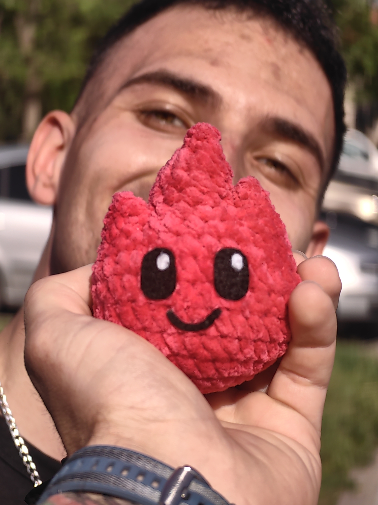 A man is holding a red, heart-shaped, plush toy with a smiling face drawn on it. The man is out of focus in the background, but his face is visible and he appears to be smiling. The man is wearing a chain. His skin is light brown and he has short, dark hair. He is wearing a watch with a blue band on his wrist. The background includes a blurred street scene with cars and trees. The specific location in Shumen, Bulgaria is not discernible from the visual data.