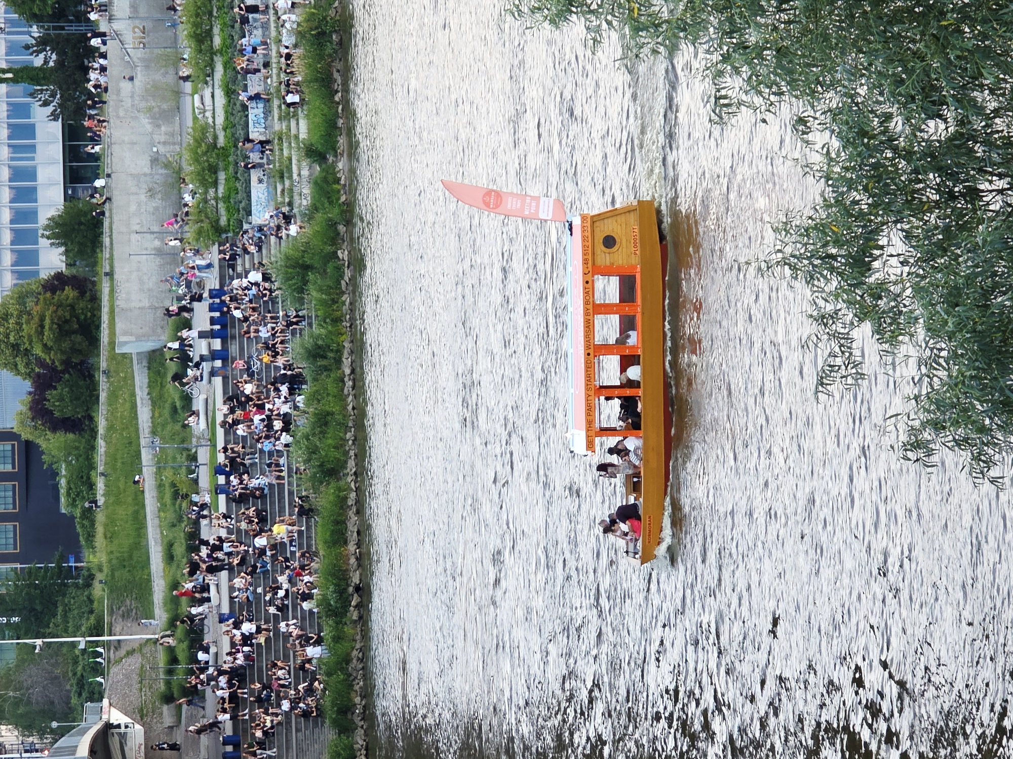 The image depicts a wide shot of the Vistula River in Warsaw, Poland, with a prominent riverboat carrying passengers. The riverboat, painted orange and red, has "GET THE PARTY STARTED! WARSAW BY BOAT" written on its side. It is moving from left to right, creating a wake. Several people are visible on the boat. In the background, the riverbank is lined with tiered concrete steps, populated by a large number of people seated and standing. Some are dressed casually, suggesting a leisure activity. Beyond the steps, there is a grassy embankment with trees and a modern building with a glass facade. The overall scene conveys a lively urban riverside atmosphere.