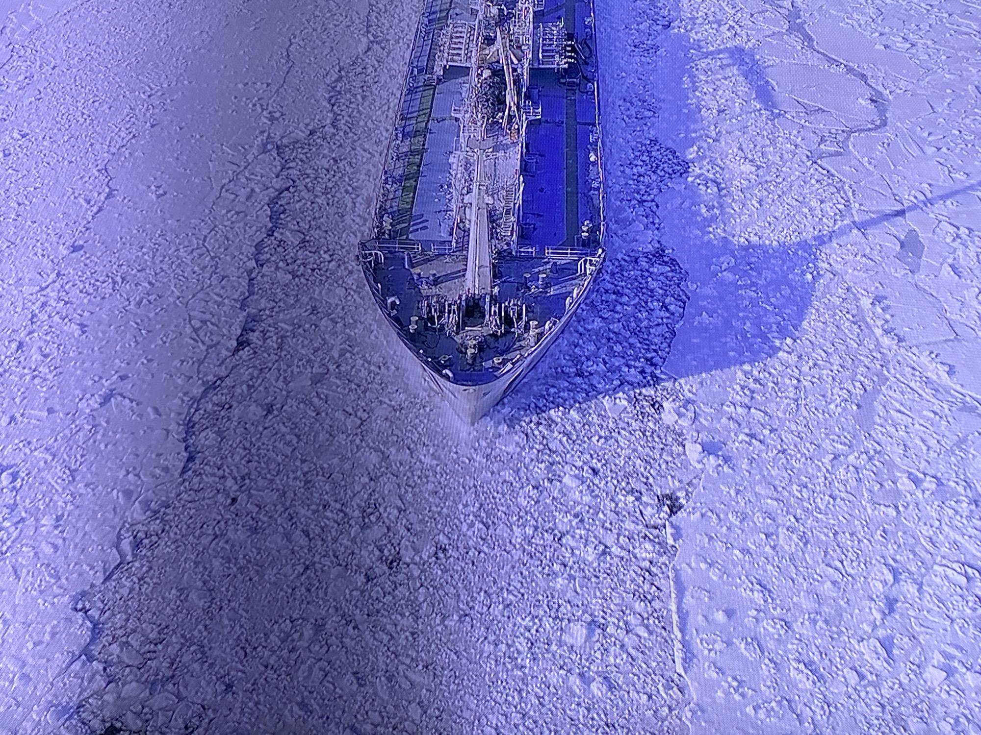 This aerial shot shows a cargo ship navigating through a frozen body of water. The water is covered in a thick layer of ice, with the ship's bow breaking through the ice. The ship itself is long and appears to be a tanker, with multiple decks and equipment visible. The surrounding ice is fragmented and varied in texture, with some areas appearing smoother and others rougher and more broken. The scene is lit by natural light, casting shadows from the ship onto the ice. The overall color palette is dominated by cool blues and purples, suggesting either a cold, possibly wintery environment or a photographic filter. The absence of any clear landmarks or distinguishing features makes it difficult to ascertain the precise location. No text is visible in the image.