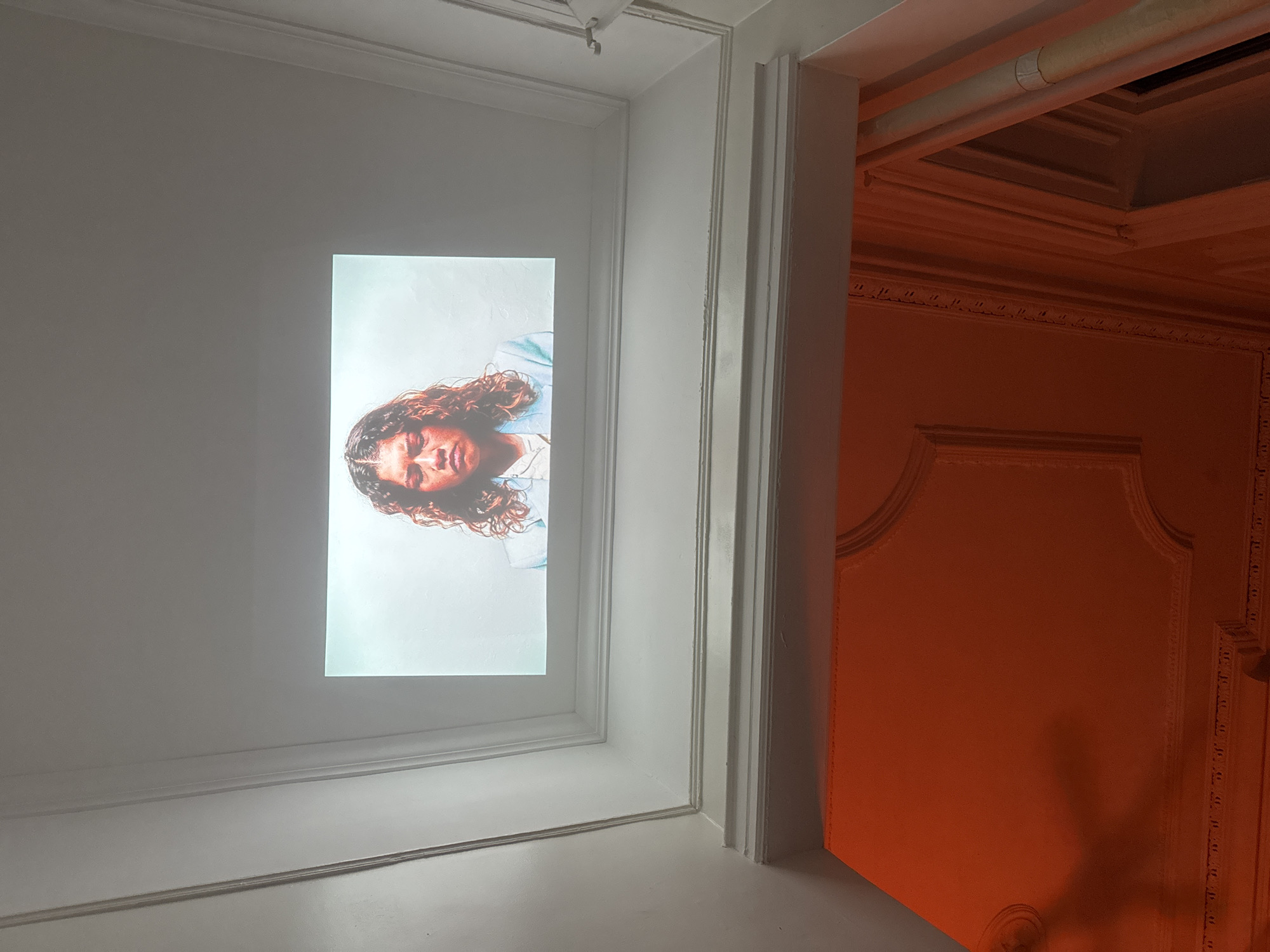 The image shows a portrait of a person projected onto a white wall. The portrait is of a person with curly hair and a light blue and white outfit. The wall is surrounded by a white frame and molding. To the right of the wall is a doorway leading to a room with orange walls and ornate molding. The environment suggests an indoor setting, possibly in a gallery or living space. Without additional context, the specific location in New York City cannot be determined.