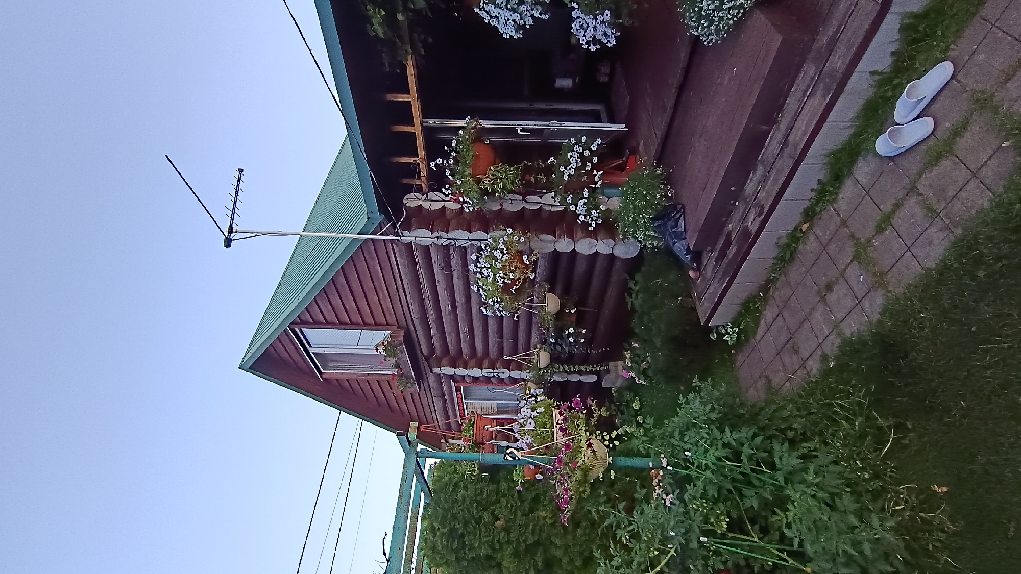 The image shows a wooden house with a green roof in a garden setting. The house is constructed of logs and has a window on the upper floor. There are several hanging flower baskets around the house, filled with white and purple flowers. A wooden porch leads to the entrance of the house, and a paved walkway is visible in front of it. A pair of light blue slippers are placed on the walkway. The garden is lush with green foliage and some smaller flowering plants. In the background, electrical wires and a TV antenna are visible against a clear blue sky.