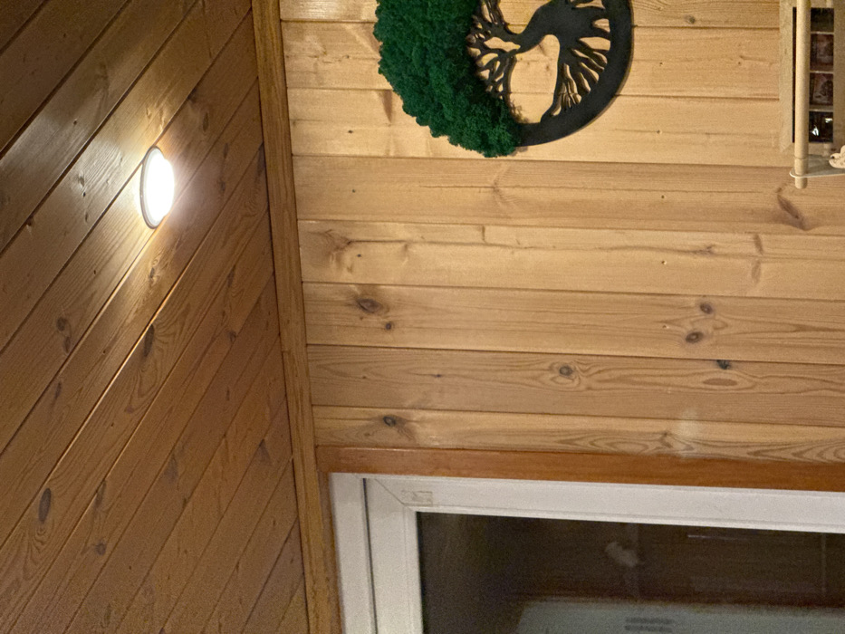 An interior scene within the city of Sysert, Russia, displays a room clad extensively in wooden paneling. The ceiling is composed of horizontal light-brown wooden planks, featuring visible wood grain and knots. A single, circular, flush-mounted LED light fixture, emitting white light, is active on the ceiling in the upper-center frame. The walls below are paneled with vertical light-brown wooden planks, also exhibiting distinct wood grain and knots. A wooden trim piece separates the ceiling and wall paneling. On the left, a portion of a white-framed doorway or window is visible, revealing a dark interior beyond, with a faint light-colored object partially discernible through the glass. On the right, a decorative wall hanging is mounted, depicting a tree with an intricate root system in a dark silhouette. The upper canopy of the tree design is filled with bright green, moss-like material. The top edge of a light-colored, multi-compartment framed object, possibly a storage unit or display holding small rectangular items, is partially visible in the lower right corner. No individuals or active subjects are present.