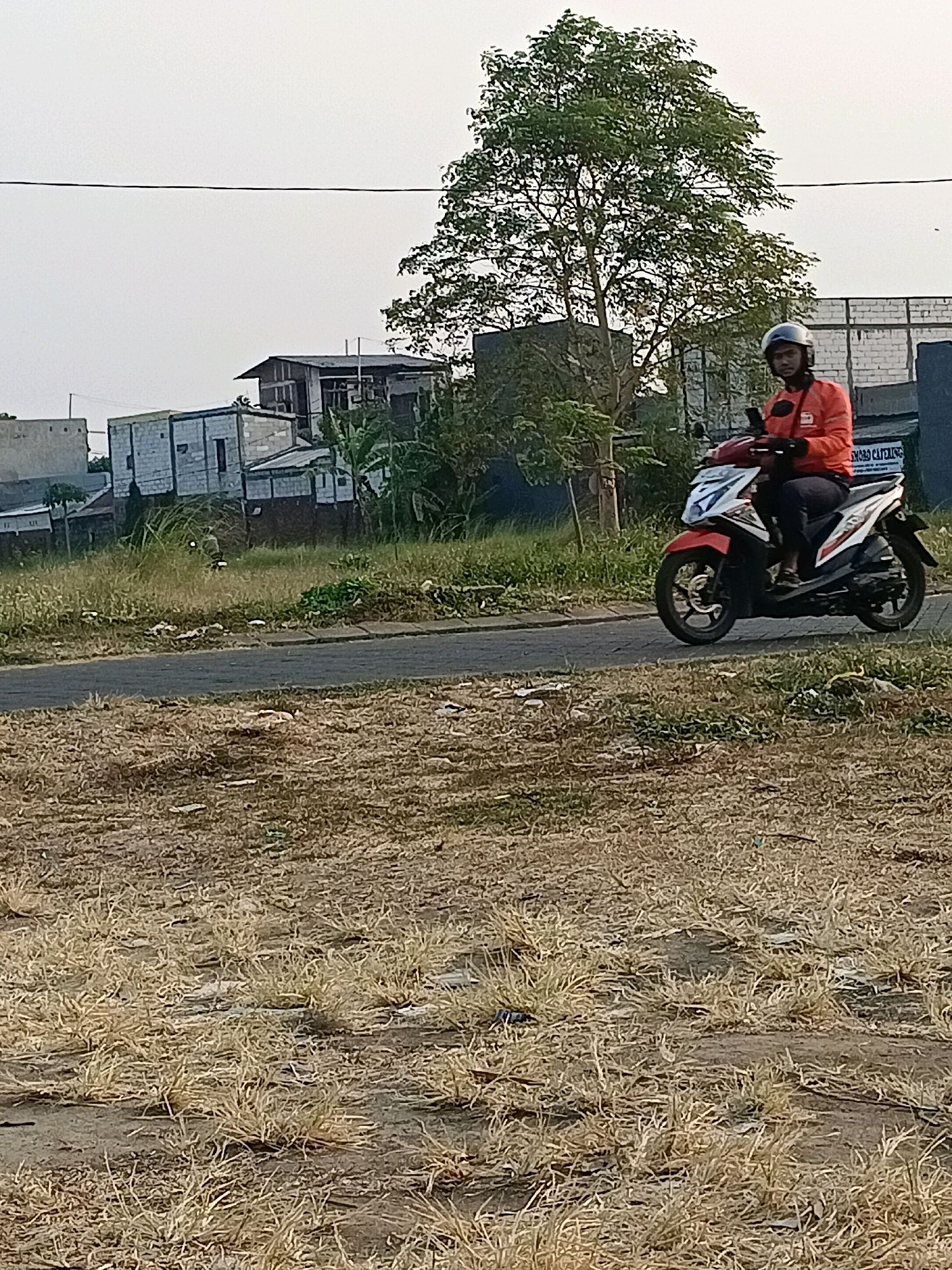A man wearing an orange jacket and a dark helmet rides a white and red motorcycle on a paved road. The man is looking towards the viewer. The motorcycle has a black seat and black wheels. The road is bordered by dry, yellowed grass and some scattered litter. In the background, there are several buildings, some with visible construction elements, and a large green tree. A white brick wall with some signage is visible to the right of the tree. The sky is overcast and a light gray color. The scene appears to be in an urban or suburban area of Tawangsari, Indonesia, based on the architecture and vegetation.