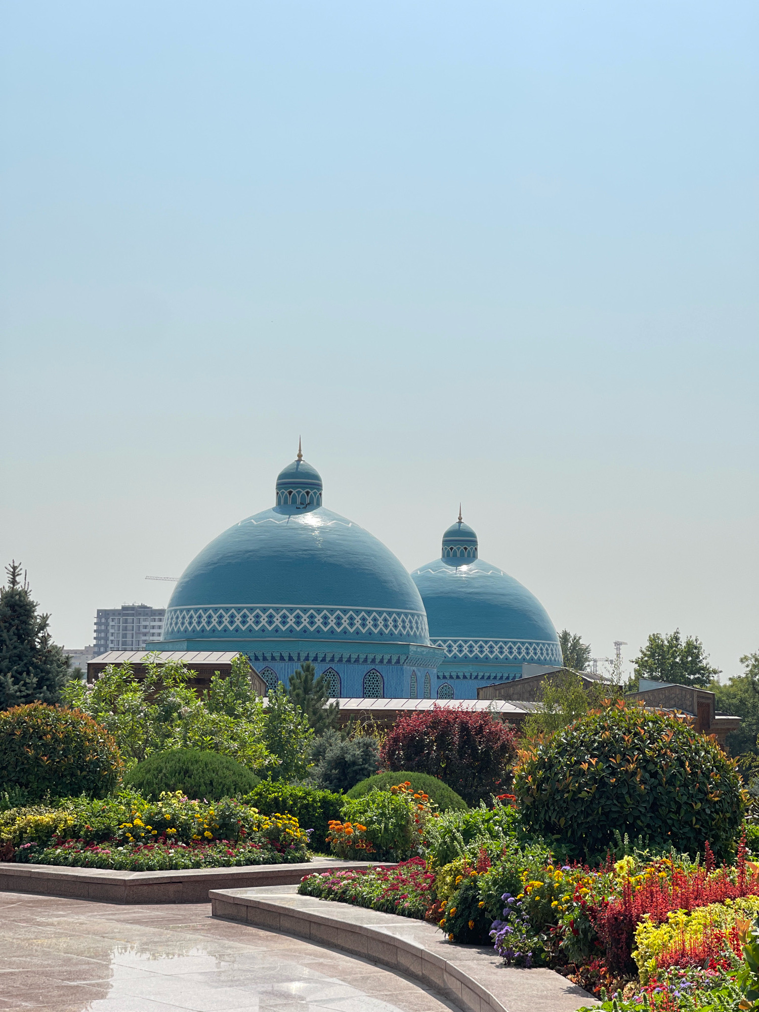 The image displays a serene outdoor scene featuring two prominent turquoise domes, characteristic of Islamic architecture, likely part of a religious complex or mausoleum. The domes are decorated with intricate geometric patterns in white. In the foreground, a vibrant and well-maintained garden bursts with a variety of colorful flowers and lush green foliage, arranged in landscaped beds bordered by stone. The paving in the immediate foreground appears to be made of polished stone, reflecting the light. Behind the domes, the urban landscape of Tashkent, Uzbekistan, is visible, with modern buildings and construction cranes suggesting ongoing development. The sky is a clear, pale blue, indicative of a sunny day. The overall impression is one of tranquility and beauty, blending architectural grandeur with natural splendor in an urban setting. This specific location is the Mausoleum of the Khazrati Imam.