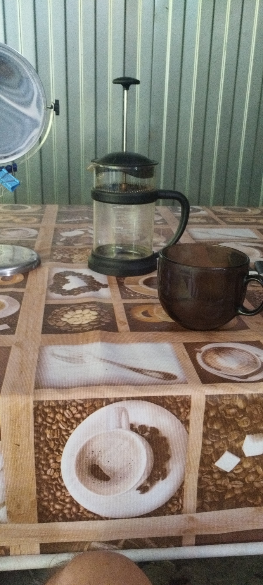 The image shows a close-up view of a table covered with a tablecloth featuring coffee-themed patterns, including images of coffee beans and cups of coffee. On the table, a glass French press coffee maker with a black lid and handle stands to the left. To the right of the French press is a dark brown glass mug. The background consists of a vertically ribbed metallic wall. To the far left, a portion of a round metallic object, possibly a lid or tray, is visible, with a blue clip attached. A person's leg is partially visible at the bottom of the frame. The scene appears to be set indoors.