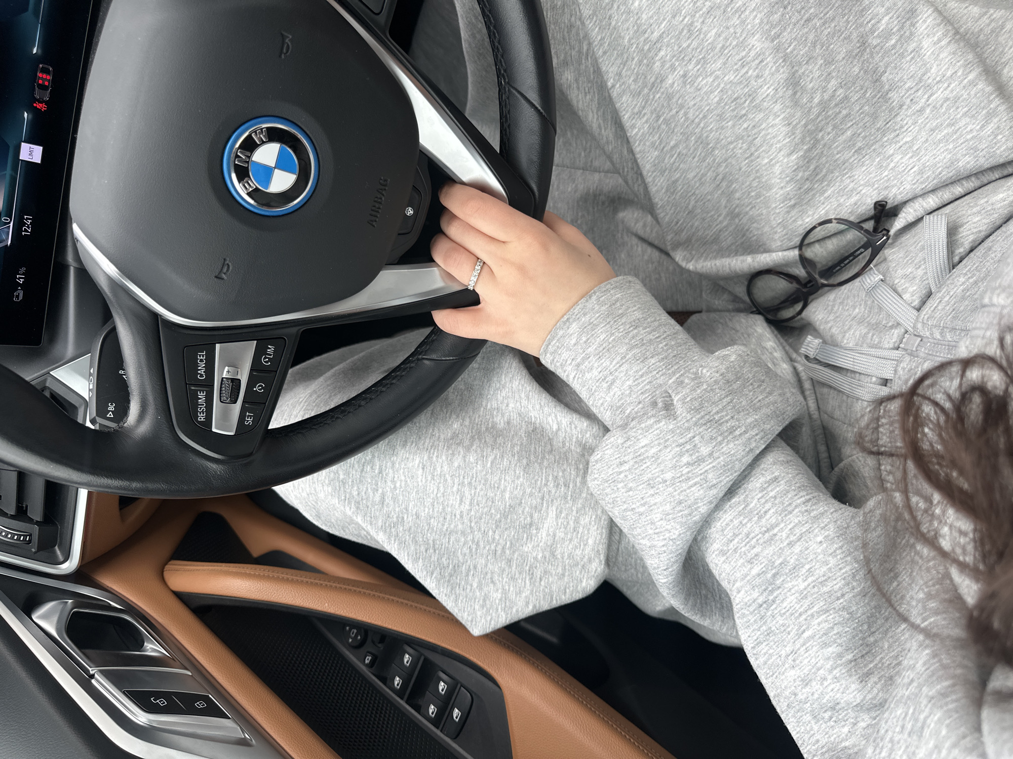 An interior view of a vehicle, specifically a BMW with electric/hybrid branding (indicated by the blue ring around the logo), is presented from the driver's perspective. The scene includes the steering wheel, a portion of the driver's body, and the driver-side door panel. A person, wearing a grey marl hooded sweatshirt and matching sweatpants, occupies the driver's seat. The individual's left hand, adorned with a diamond eternity ring on the ring finger, rests on the lower right spoke of the steering wheel. Dark brown hair is visible in the lower-left corner. A pair of tortoiseshell-framed eyeglasses lies on the driver's lap. The car's dashboard displays a time of 12:41, a 41% battery charge, a "LIMIT" warning, and an unbuckled seatbelt indicator for two rear passengers. The door panel features brown leather upholstery, a silver door handle assembly, and window controls. No external environmental details are discernible to determine a specific location within Yerevan, Armenia.