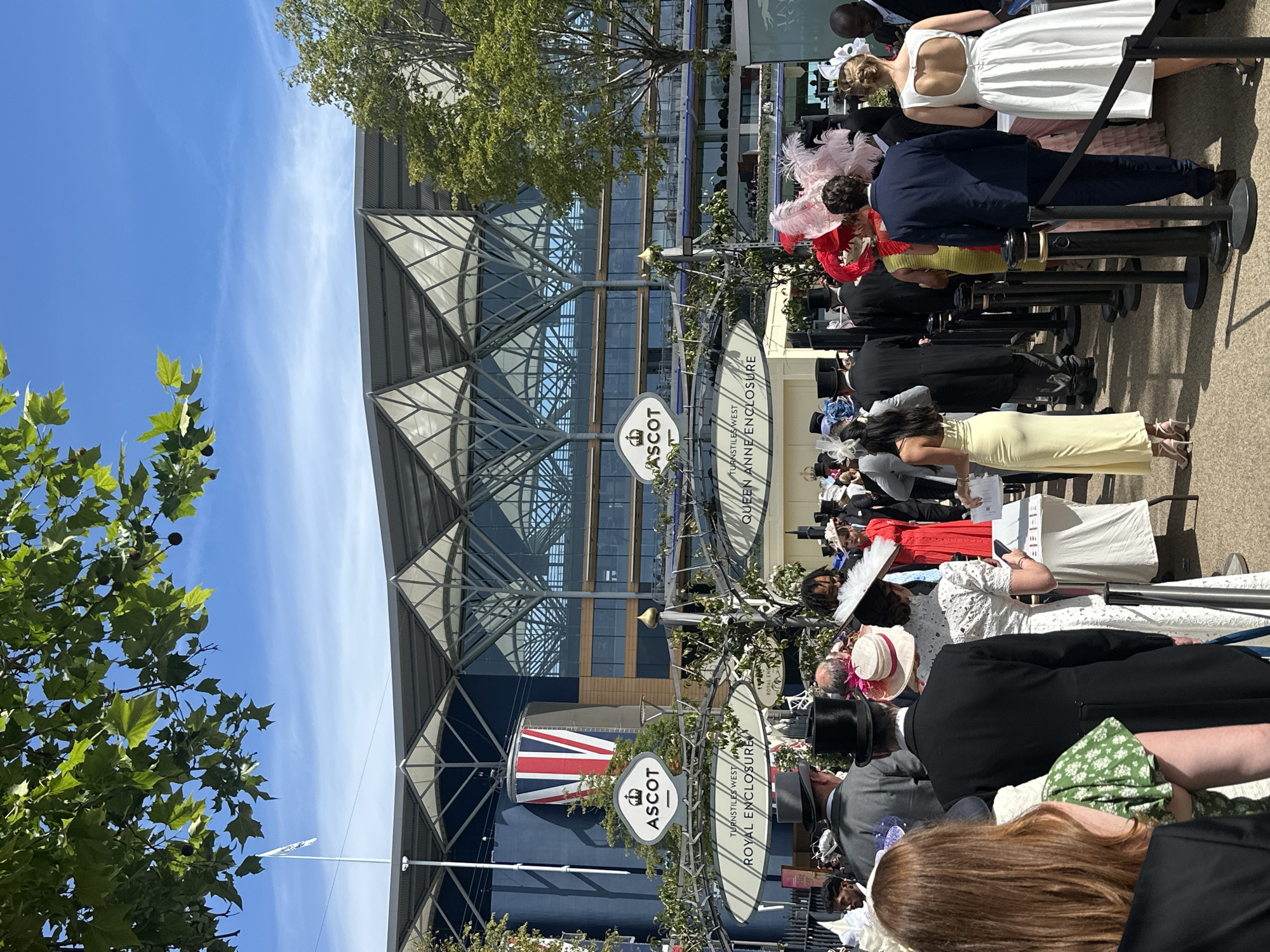 A full shot captures a crowd of people dressed in formal attire at Ascot Racecourse in Ascot, United Kingdom. The day is bright and sunny, with a clear blue sky visible above a large, modern building with a distinctive roof structure. The building features a large Union Jack flag displayed prominently on its exterior.
The foreground is filled with people queuing, many of whom are wearing elaborate hats and formal wear, characteristic of events like Royal Ascot. Men are seen in top hats and tailcoats, while women are in elegant dresses and striking hats adorned with feathers and flowers. The crowd appears to be moving in a line, guided by black stanchions.
Signage visible in the image indicates "ASCOT," "TURNSTILES WEST," and "ROYAL ENCLOSURE," confirming the location and specific area within the racecourse. Lush green foliage from trees overhangs parts of the scene, adding a touch of nature to the urban setting. The overall atmosphere suggests a high-profile event where spectators are dressed for a special occasion.