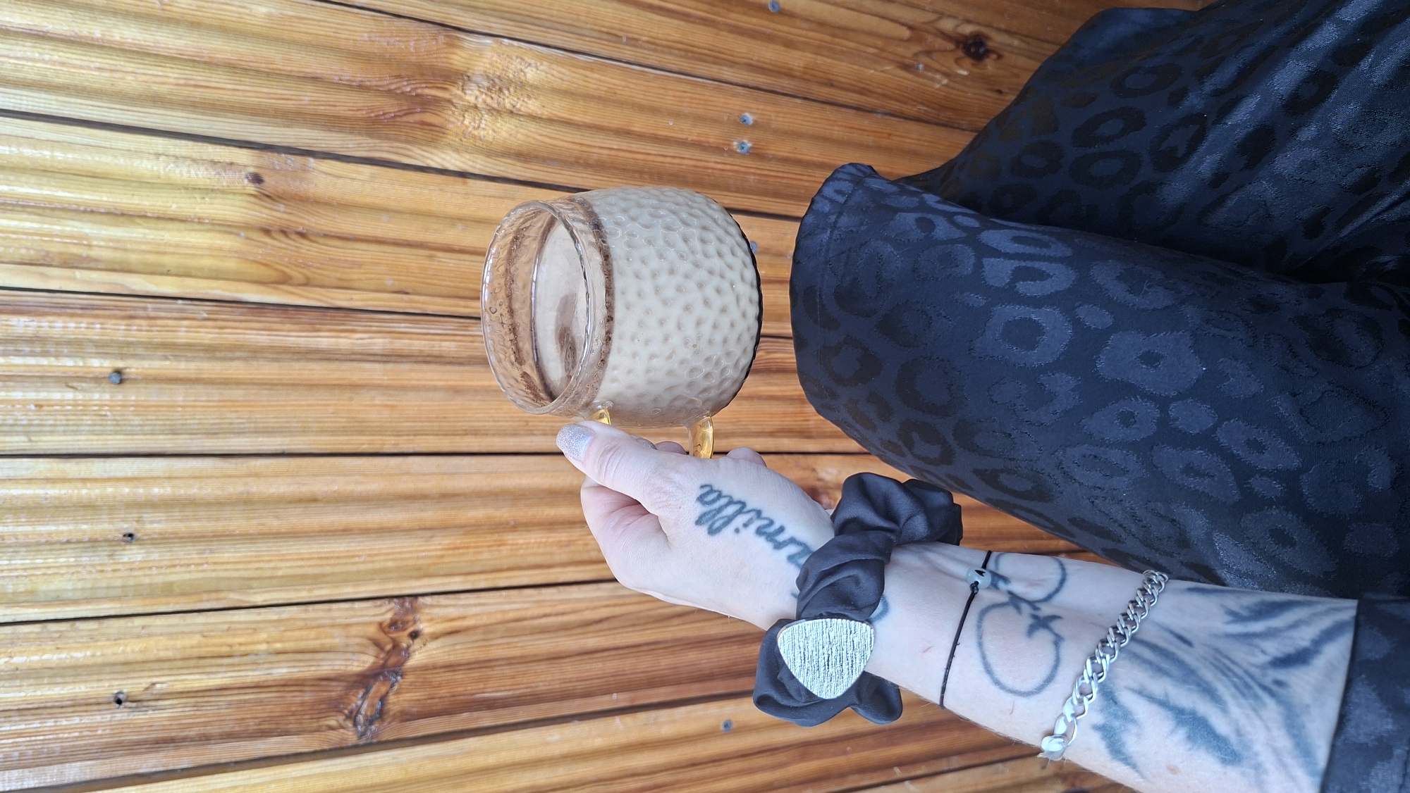 The image features a person's hand holding a glass jar filled with a light brown liquid, possibly honey or a beverage. The jar has a textured, dimpled surface and a shiny rim. The background is composed of wooden planks, laid horizontally, suggesting an outdoor or rustic setting. To the right of the jar, a black fabric with a subtle leopard print pattern is visible, part of clothing or an accessory.
The person's hand has several tattoos, including the word "camilla" written in cursive on the back of the hand and a design on the wrist. They are wearing a black scrunchie with a triangular, textured silver charm and a silver chain bracelet. The lighting suggests it's daytime, with natural light illuminating the scene. The overall mood appears casual and relaxed.
No specific event or time of day can be definitively determined. The location context provided is Забалоцкі сельскі Савет, Belarus.