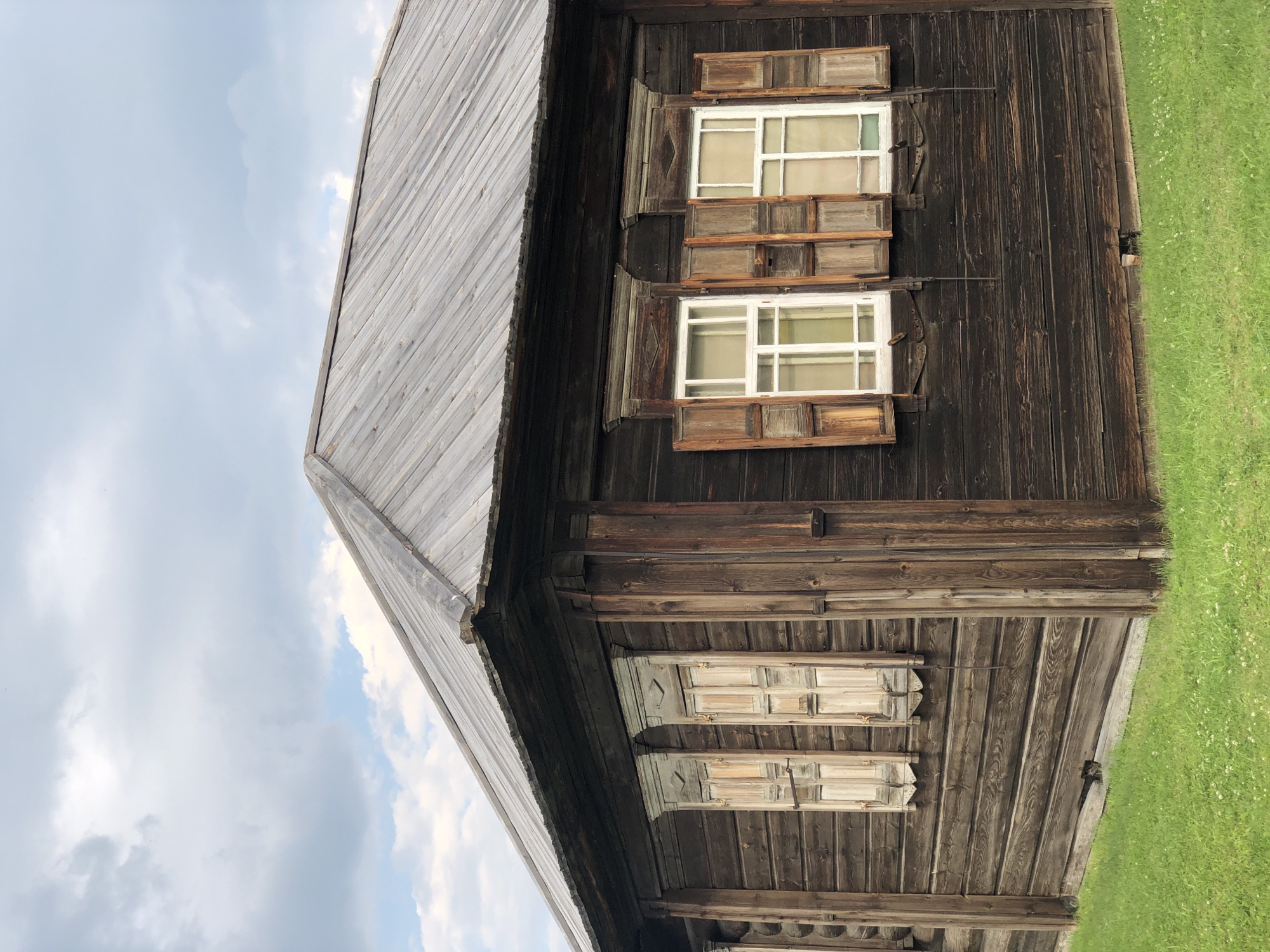 The image shows a corner view of a dark wooden log house with a metal roof, located in Shushenskoye, Russia. There are four windows visible, each with weathered wooden shutters. Two windows on the left have their shutters closed, while the two windows on the right have their shutters partially open, revealing panes of glass. The house is situated on a patch of green grass. The sky above is cloudy with patches of blue.