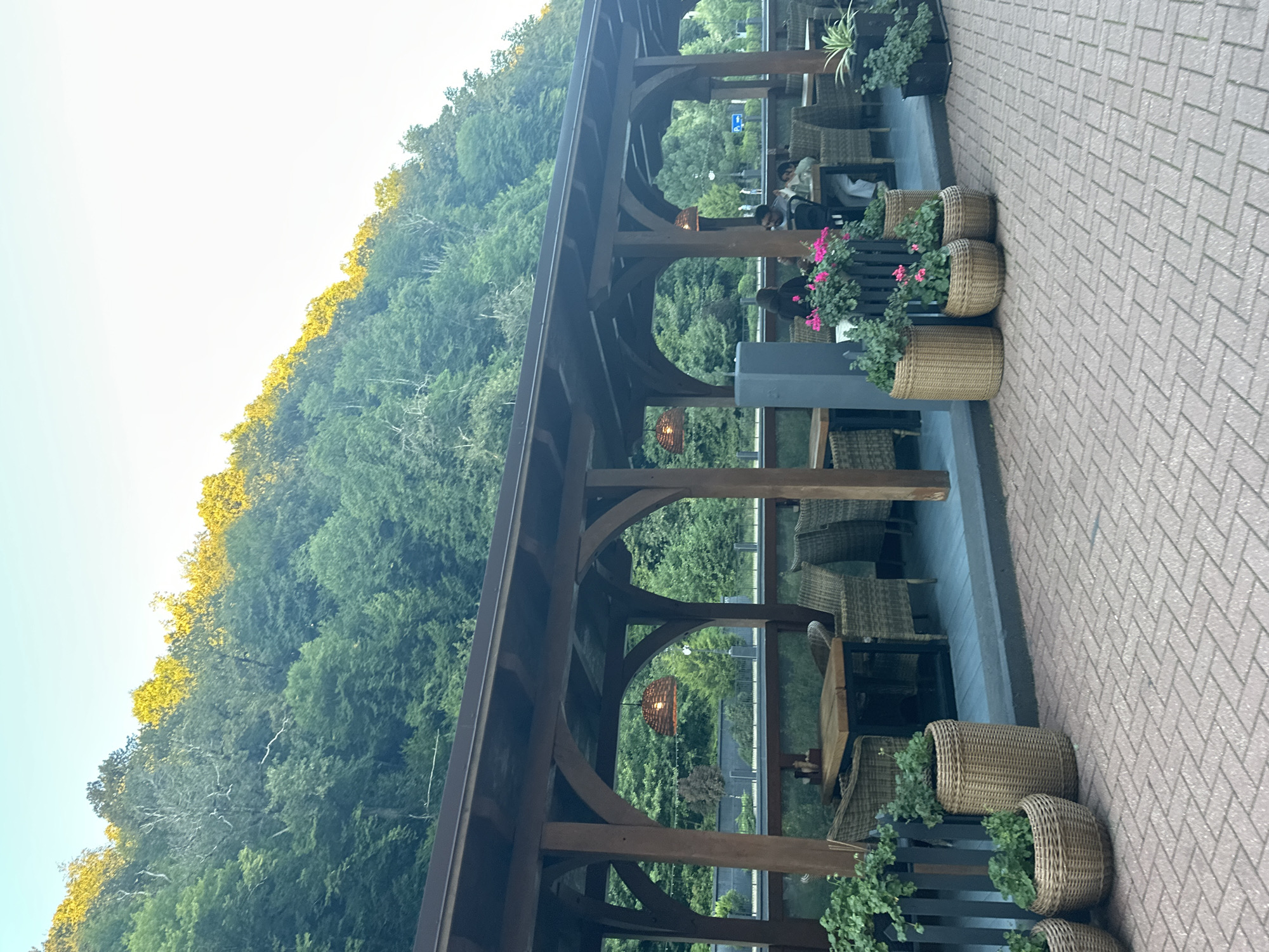 The image captures an outdoor seating area of a restaurant in Estosadok, Russia, nestled at the base of a lush, tree-covered mountainside. The sun casts a warm, golden glow on the upper canopy of the trees, creating a scenic backdrop. The seating area is covered by a dark wooden pergola with a sloped roof. Several tables and chairs, made of woven wicker and wood, are arranged under the pergola, some occupied by patrons. The floor of the seating area appears to be a dark blue composite material, while the exterior walkway is paved with reddish-brown brick pavers arranged in a herringbone pattern. Various potted plants, including flowering ones and greenery, are placed around the seating area and along the walkway, adding to the ambiance. Two wicker lamps hang from the pergola, casting a soft light. In the background, beyond the seating area, there are more trees and the suggestion of a road with some traffic. A few people are visible sitting at tables under the pergola, engaged in conversation or dining.
