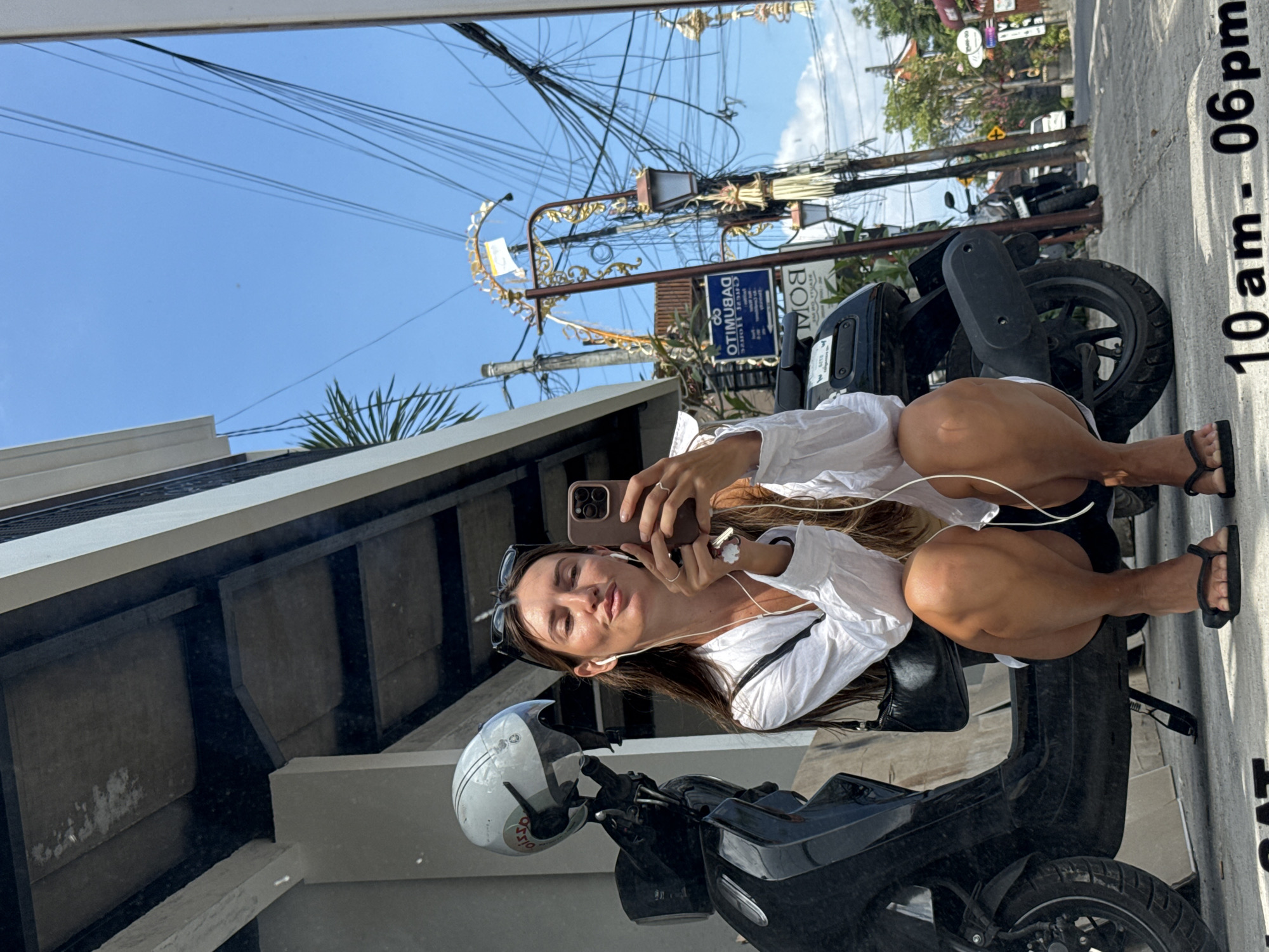 A woman with long brown hair and sunglasses perched on her head takes a mirror selfie while sitting on a scooter in North Kuta, Indonesia. She is wearing a white button-up shirt, black shorts, and black flip-flops. She has white earbuds in and is holding a smartphone in her right hand, appearing to be taking the photo. The scooter is black with a white helmet on the handlebars. In the background, there are buildings, power lines, streetlights, and other scooters. A sign with text is visible on a pole. The time "10 am - 06 pm" is superimposed on the lower portion of the image.