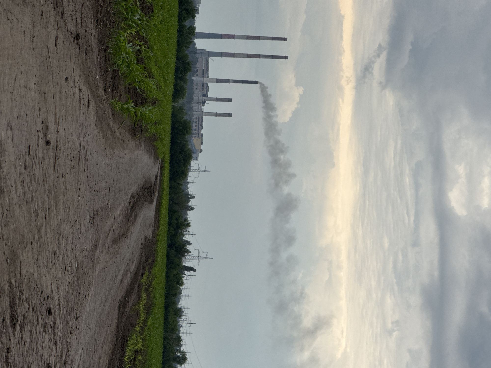 The image presents a landscape juxtaposing agricultural land with an industrial complex, set under an overcast sky in Zanky, Ukraine. In the foreground, a textured, light brown dirt path, appearing somewhat damp, curves gently and recedes into the distance. This path is bordered by a vibrant green field, likely cultivated crops, which forms a dense line across the lower left and center of the frame.
Behind the green field, a line of darker green trees separates the foreground from the industrial elements in the midground and background. The industrial complex is dominated by several tall, slender chimneys, painted with alternating white and red bands. One of these chimneys is actively releasing a thick, dark grey plume of smoke that rises into the sky, indicating ongoing operations and industrial activity. Various other structures of the industrial plant are visible behind the chimneys. Further across the landscape, a series of electricity transmission towers (pylons) can be seen.
The sky is largely overcast, filled with varying shades of grey and white clouds. A distinct, brighter vertical band of light cuts through the clouds on the right side of the image, suggesting either diffused sunlight breaking through heavy cloud cover or the light of an early morning or late afternoon sun. There are no people visible in the image, nor is any text present. The scene effectively captures the coexistence and contrast between nature and heavy industry.