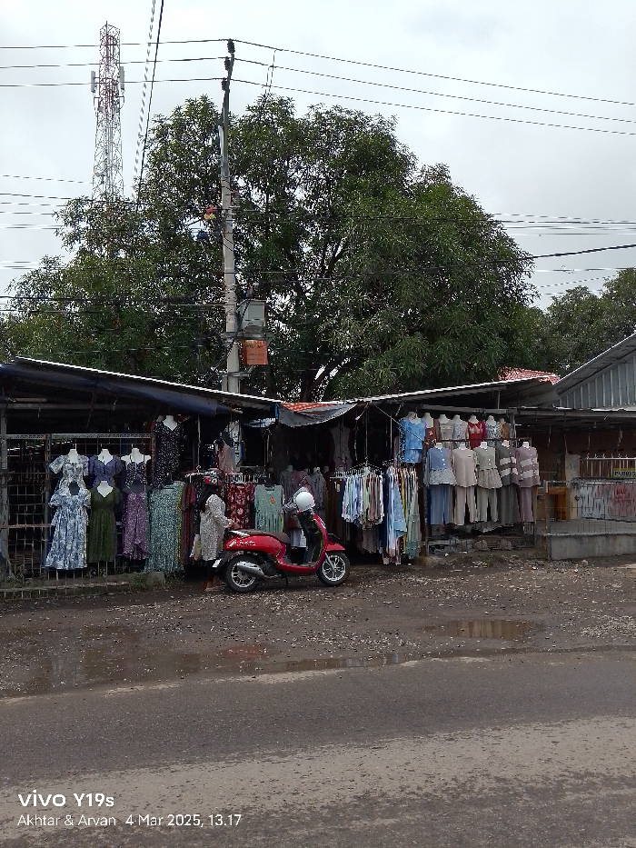 The image shows a street market in Jatibarang, Indonesia, located at coordinates -6.466, 108.299. The market consists of several stalls with clothes hanging on display. A red scooter is parked in front of the stalls, driven by a person wearing a white helmet. The environment is urban, with a paved road and a telephone pole with wires running across it. The ground is wet and there is a puddle in front of the stalls. There are a few trees behind the stalls, and a building in the background. There is a large telecommunications tower visible in the background. The sky is overcast, indicating a possible rainy day. The image was captured by a person who is standing outside the market, facing the stalls. The image was taken on a mobile device and there is a timestamp indicating the date and time the image was taken.