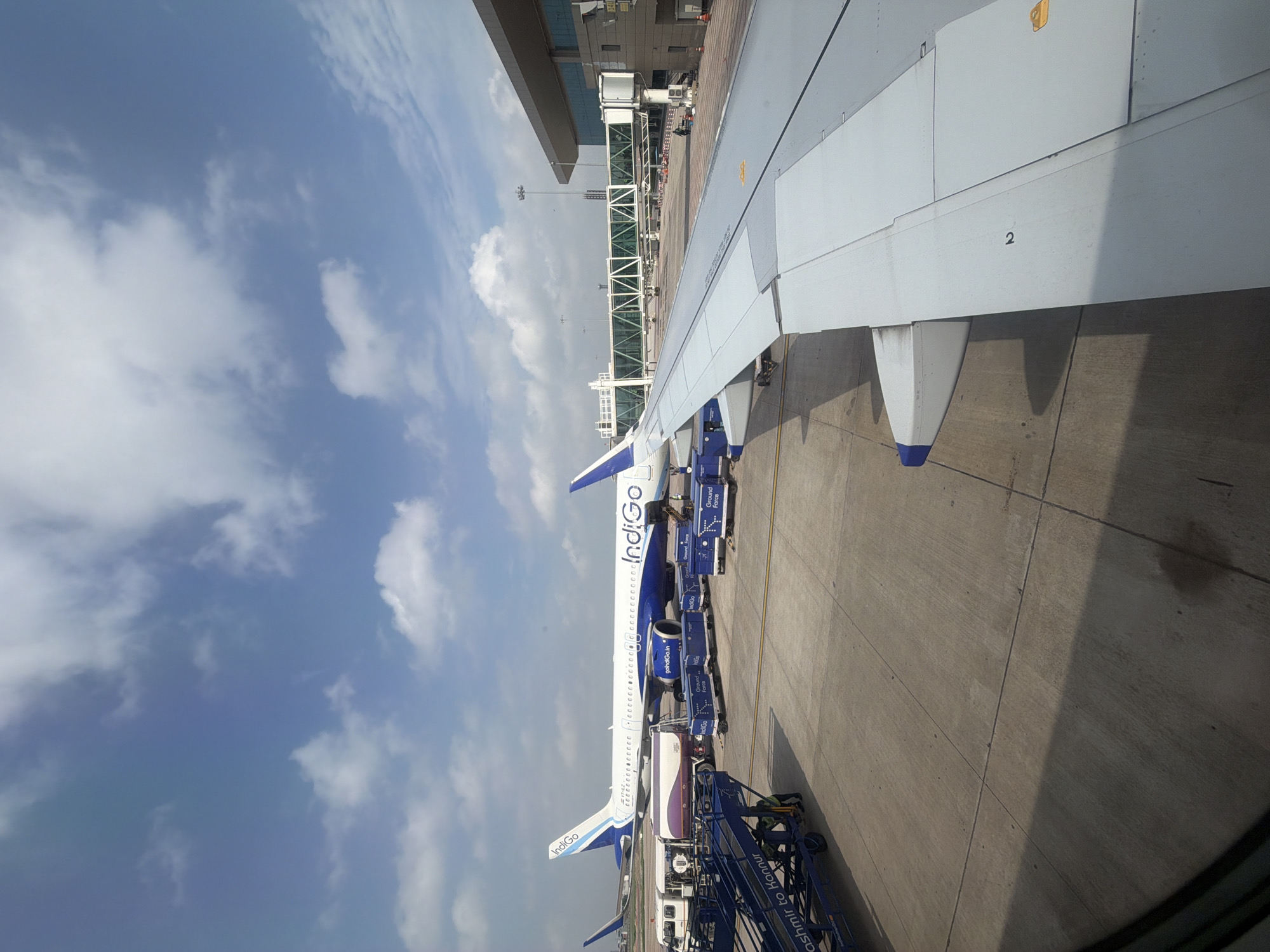 An aerial view captures an Indigo airline plane parked on the tarmac at an airport, possibly in the vicinity of Vikram Nagar, India, under a partly cloudy blue sky. The aircraft, predominantly white with blue accents, is labeled with "Indigo" on its fuselage and tail. Several ground support vehicles and equipment, including luggage carts and a fuel truck, are positioned around the plane. A jet bridge is connected to the terminal building, which is visible in the background. The foreground is dominated by the wing of another aircraft, suggesting the perspective is from inside a plane. The tarmac is paved concrete, marked with various lines and shadows cast by the aircraft and structures. No specific actions or interactions are clearly discernible, but the scene implies standard airport operations.
