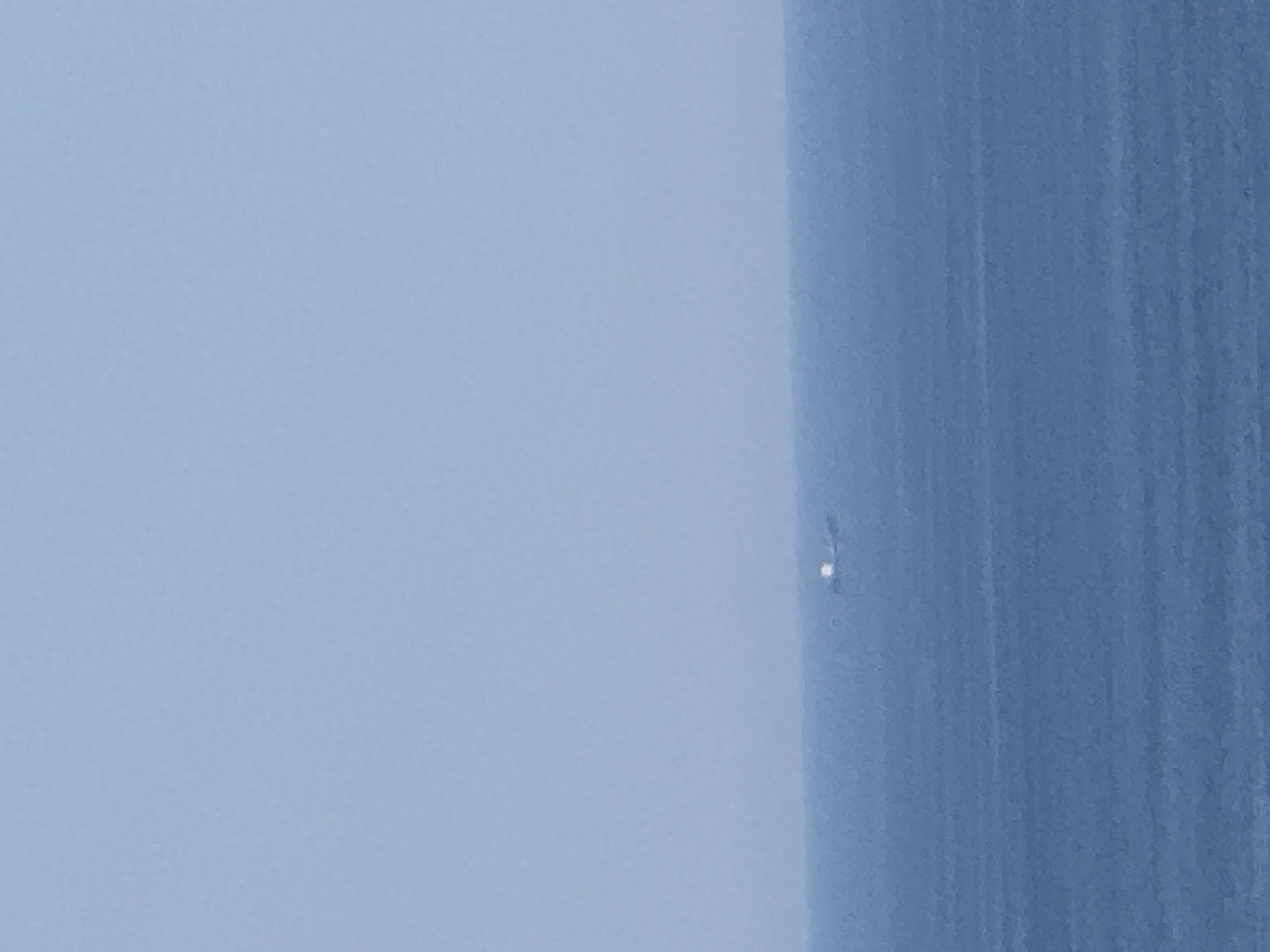 The image displays a minimalist, abstract composition with a strong emphasis on color and texture. The left two-thirds of the frame are dominated by a soft, light blue gradient, suggesting a hazy or atmospheric sky. The texture in this area appears slightly grainy, as if captured on film or with a subtle noise filter.
The right third of the image features a vertical band of a deeper, richer blue. This band exhibits a distinct textured pattern, resembling vertical brushstrokes or the grain of a weathered surface. The contrast between the smooth gradient on the left and the textured band on the right creates a sense of depth and visual interest.
Near the center of the image, on the boundary between the light blue and the darker textured band, is a small, bright white speck. This speck is the only distinct "object" in the frame, and its isolated nature draws the viewer's attention. Its origin is unclear and could be a lens flare, a dust particle, or a very distant light source.
The overall impression is one of quietude and simplicity. The colors are serene, and the abstract nature of the composition invites interpretation. There is no discernible activity, time of day, or specific location cues beyond the abstract color and texture. The image feels like a study of light and form, possibly capturing a moment of atmospheric diffusion or a close-up of a textured surface against a blurred background. No text is visible in the image.