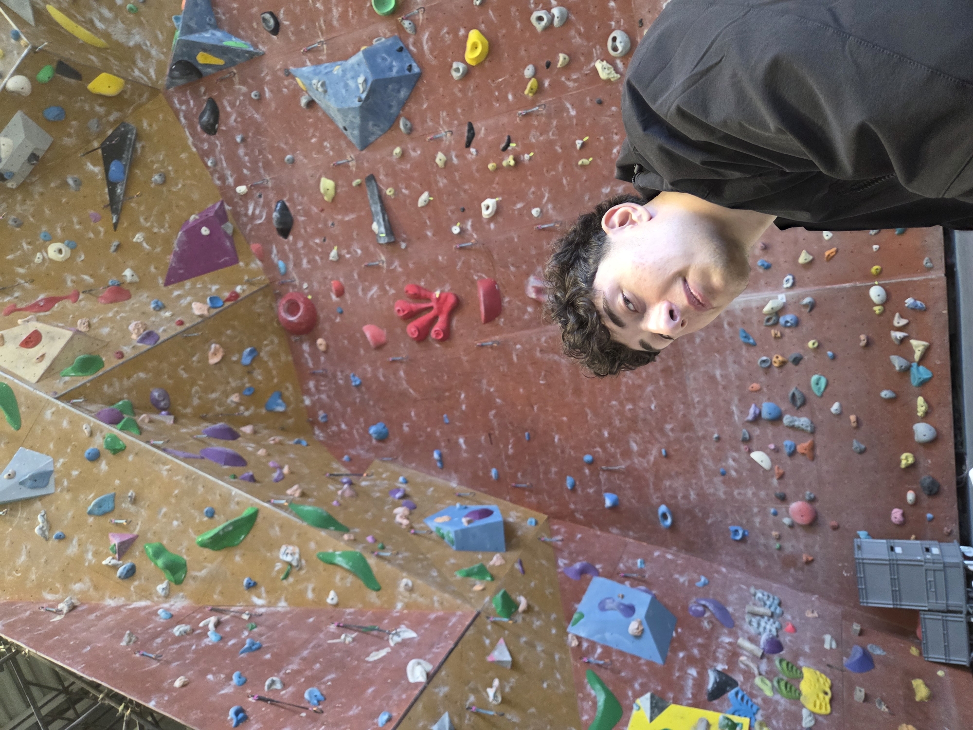The image displays a young male situated in an indoor bouldering gym in Sofia, Bulgaria. The subject, a male in his late teens or early twenties with short, dark curly hair and a fair complexion, occupies the lower right portion of the frame, wearing a dark jacket and looking directly at the viewer with a neutral expression. The background consists of a large, multi-faceted climbing wall, featuring sections in reddish-brown and tan colors. This wall is densely covered with numerous climbing holds of various shapes, sizes, and colors, including green, blue, yellow, red, purple, black, white, and grey, along with several large, angular climbing volumes. In the lower-left background, three gray plastic crates are stacked. Visible in the upper-left corner are structural elements consistent with an industrial building's ceiling. The scene is illuminated by consistent artificial lighting.