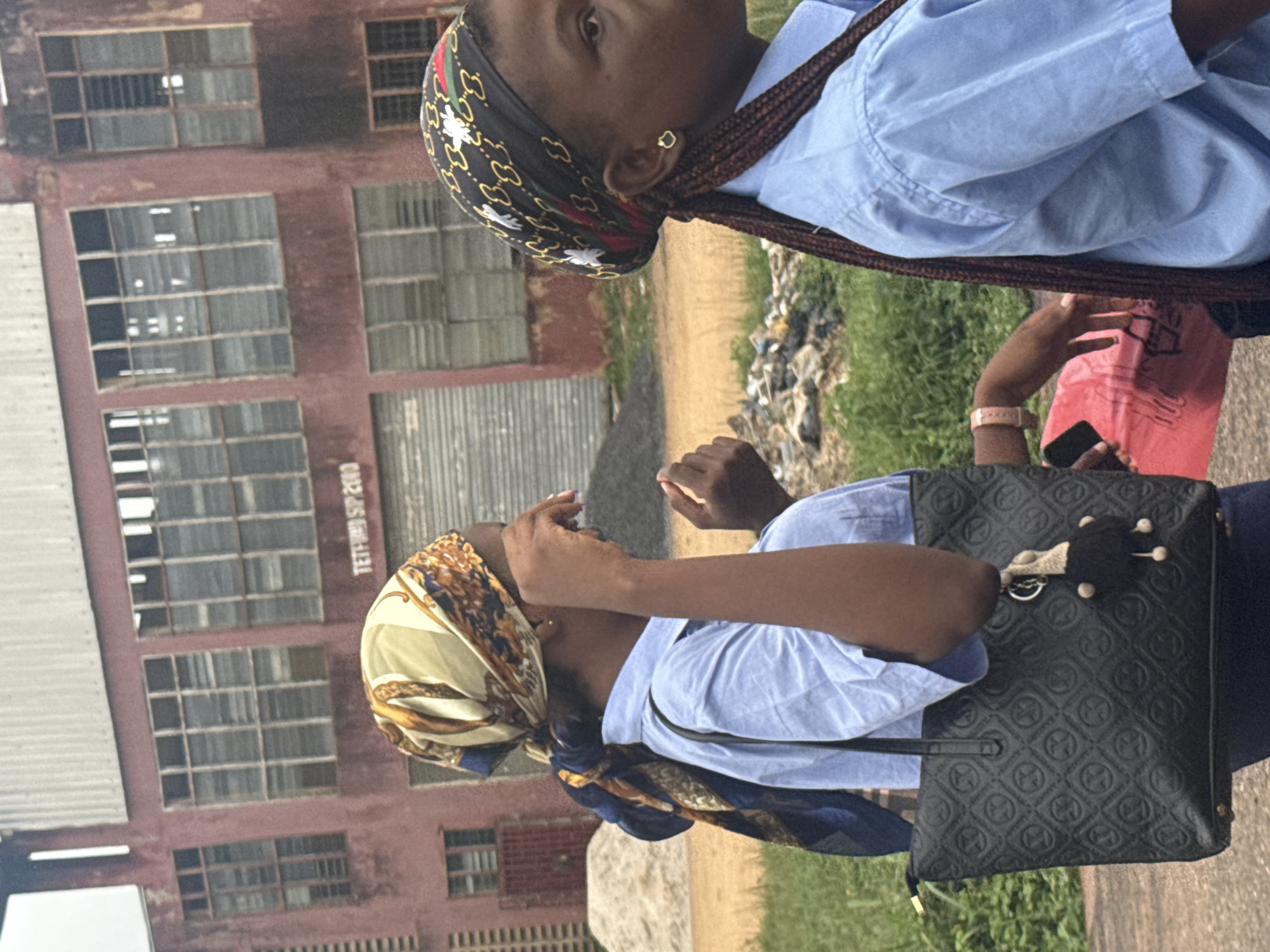 In the city of Ihiagwa, Nigeria, two young females, likely students, are captured in a medium shot, standing in front of a large, aged, reddish-brown building with numerous windows. The building, possibly an industrial or institutional structure, has a corrugated metal roof. The first female, on the left, wears a light blue collared shirt and a patterned headscarf with a blend of cream, blue, and gold. She carries a large, textured black handbag with a decorative keychain. Her right hand is raised near her face, and her left hand is not fully visible. The second female, to her right, is partially obscured by the first. She also wears a light blue collared shirt and a black headscarf adorned with a repetitive white pattern and a red and green trim. Her braided hair is visible, and she wears a small, heart-shaped earring. Both females appear to be walking or standing. The environment includes green grass and dirt in the foreground, with piles of rubble or construction material to the left of the building. A red plastic bag is held by someone or something just out of frame to the right, with a hand visible reaching towards it. The lighting suggests it is daytime.
