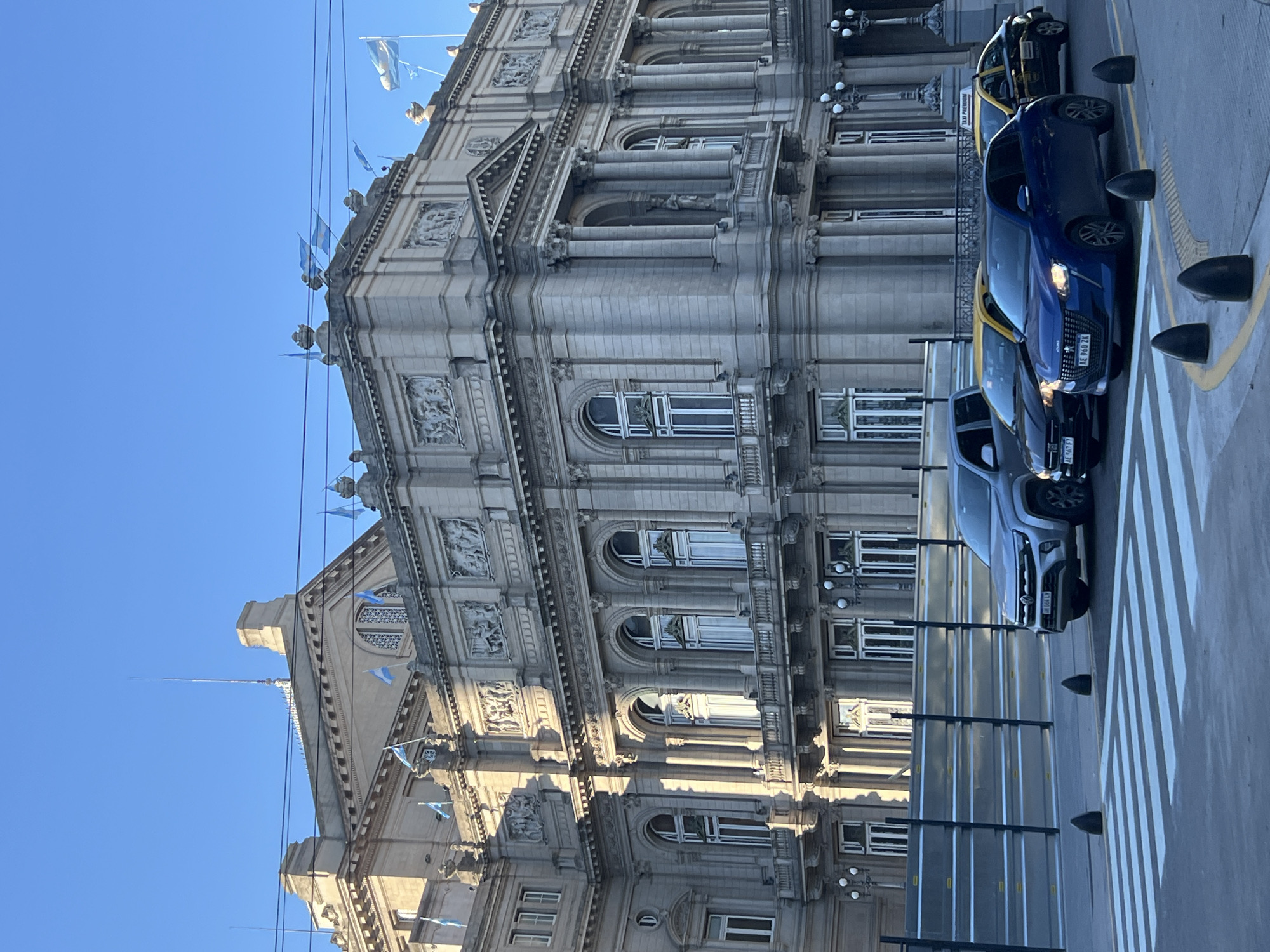 The image depicts the Palacio de Correos in Buenos Aires, Argentina. The building has a neo-classical architecture with decorative facades, columns, arched windows, and carvings. Several Argentinian flags are displayed on the top of the building. Parked cars, including taxis, are on the side of the street. A crosswalk is visible on the street in front of the building. The sky is clear and blue.