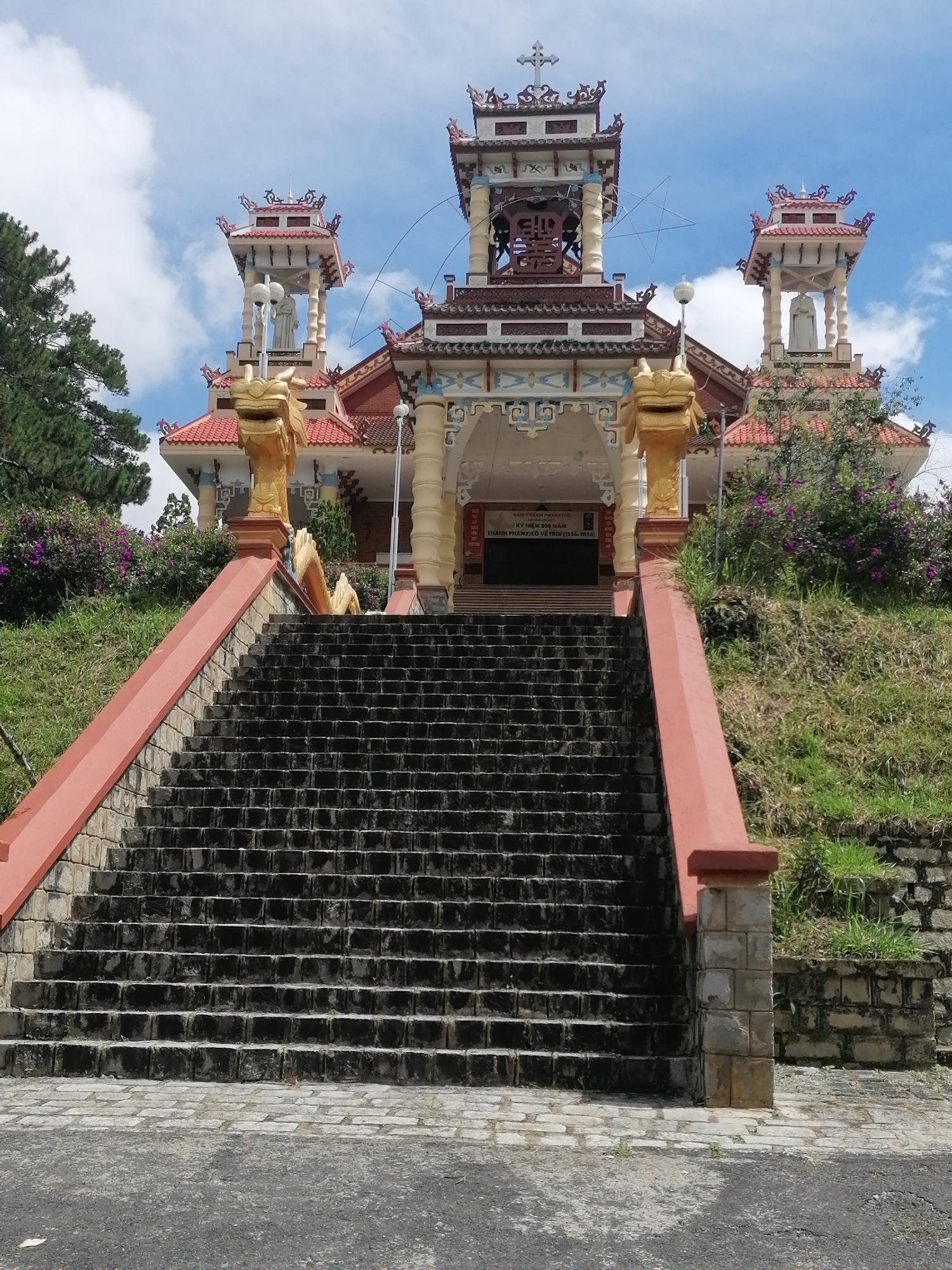 The image shows a wide, dark stone staircase leading up to a religious building with traditional Vietnamese architecture. The building features ornate roofs with upturned eaves, decorative motifs, and towers topped with crosses. Golden dragon sculptures are prominently placed on either side of the entrance. Green grass and flowering bushes flank the staircase, and the sky above is partly cloudy. Based on the architectural style and the presence of a sign with Vietnamese text, this building is likely a Catholic church or monastery in Da Lat, Vietnam. The scene is captured from a low angle, emphasizing the scale of the staircase and the grandeur of the building. No people are visible in the image.