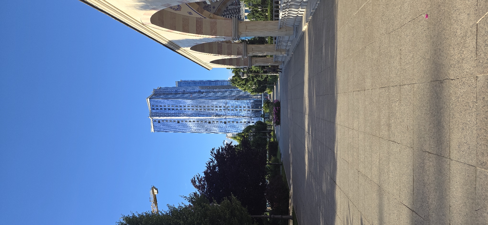 The image presents a view of Grozny, Russia. Prominently visible are modern high-rise buildings with reflective glass facades, set against a clear blue sky. In the foreground, an arched structure, possibly part of a historical or cultural site, borders a wide, paved walkway. The pathway, under sunlight, casts long shadows, indicating the time of day is likely early morning or late afternoon. Trees line the walkway, adding a touch of greenery to the urban landscape. A construction crane is seen in the distance. There is no evidence of people or actions.