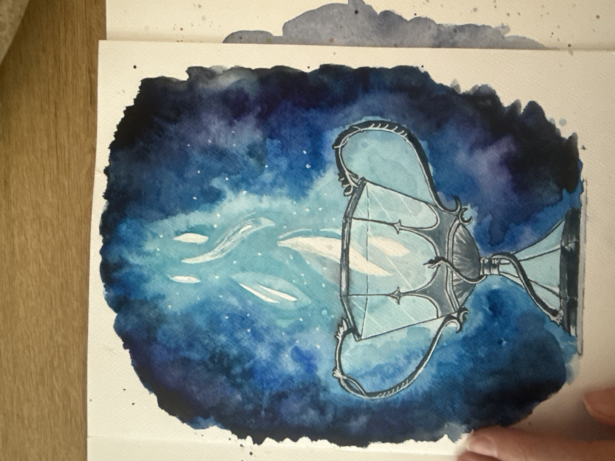 The image is a watercolor painting of a goblet surrounded by a dark blue, starry background. The goblet is depicted with intricate details, appearing to be made of glass or crystal, with a silvery, ornate base and handles. Flames, rendered in white and light blue watercolor, emanate from the goblet, creating a magical or ethereal effect. The background is a wash of deep blues and purples, speckled with white dots to represent stars, fading out to the edges of the paper. The painting is on a white paper surface, which is placed on a wooden surface. There are no people or specific landmarks visible in the painting, thus its location within the city of Wrocław, Poland, cannot be determined.