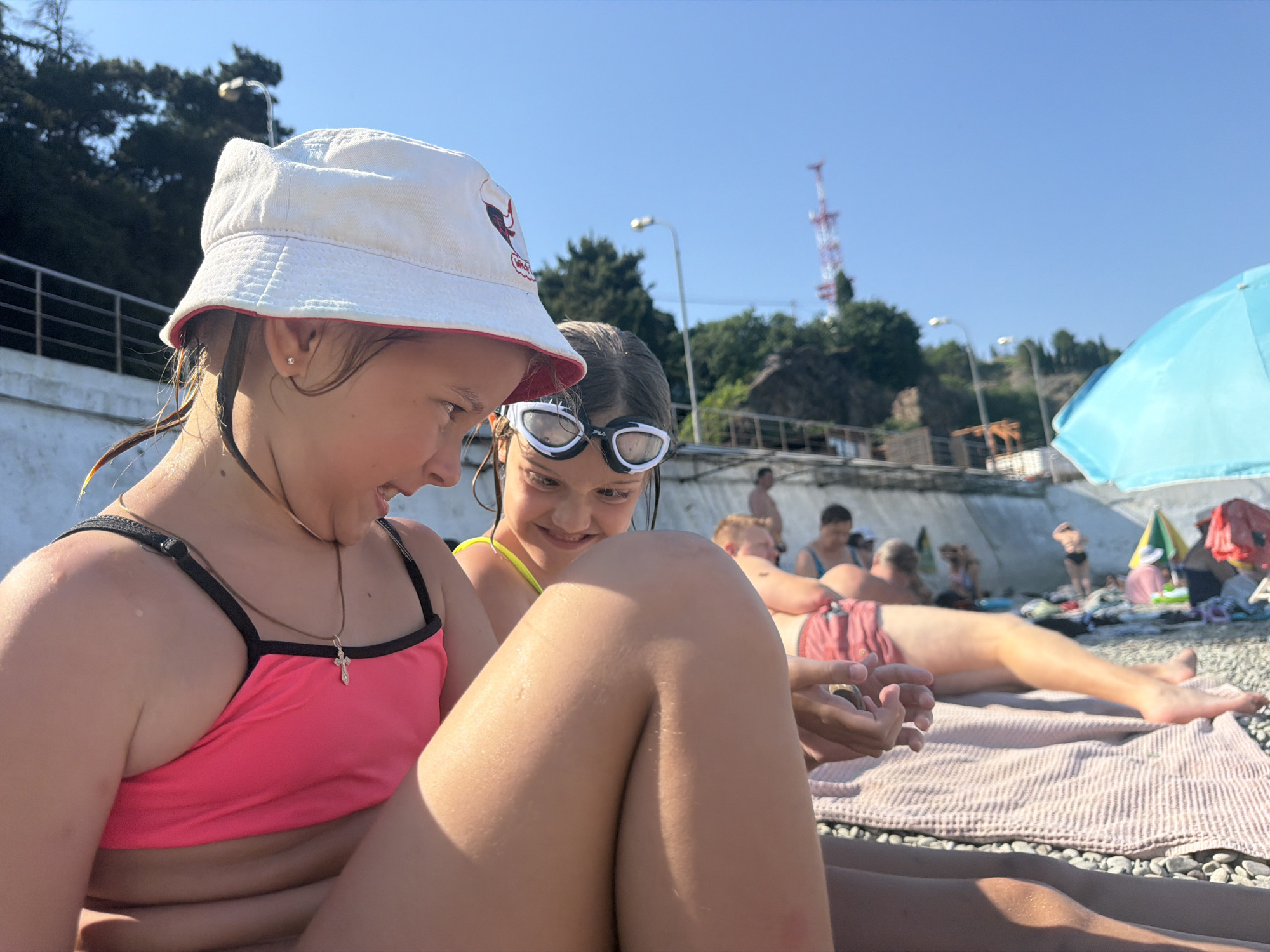 This is a close-up, eye-level medium shot of two young girls on a pebble beach, likely during a sunny day. The girl in the foreground, wearing a pink bikini top and a white bucket hat with a red trim, is looking down and to her left with a slight smile. Her wet hair is visible. She is wearing a cross pendant necklace. Beside her, to her right, is another girl wearing swimming goggles and a yellow bikini top. She is also looking down, and her expression suggests amusement. Her eyes are barely visible behind the goggles.
The background shows a beach scene with other people relaxing. A man in red patterned swim trunks is lying on a striped towel in the middle ground. Further back, several other people are visible, some sitting and some standing. Beach umbrellas, one a bright blue, are dotted around the scene. In the far background, trees and a concrete wall suggest a hillside or cliff face. A tall, red and white tower, possibly a communication tower, is visible on the hillside, and a street lamp is also present.
The bright sunlight and clear blue sky indicate it's daytime, likely late morning or afternoon, and the weather is warm and clear. The setting is a beach, with pebbles visible in the foreground and on the ground where people are lying. The overall mood appears relaxed and happy, consistent with a beach vacation. No visible text is discernible in the image.