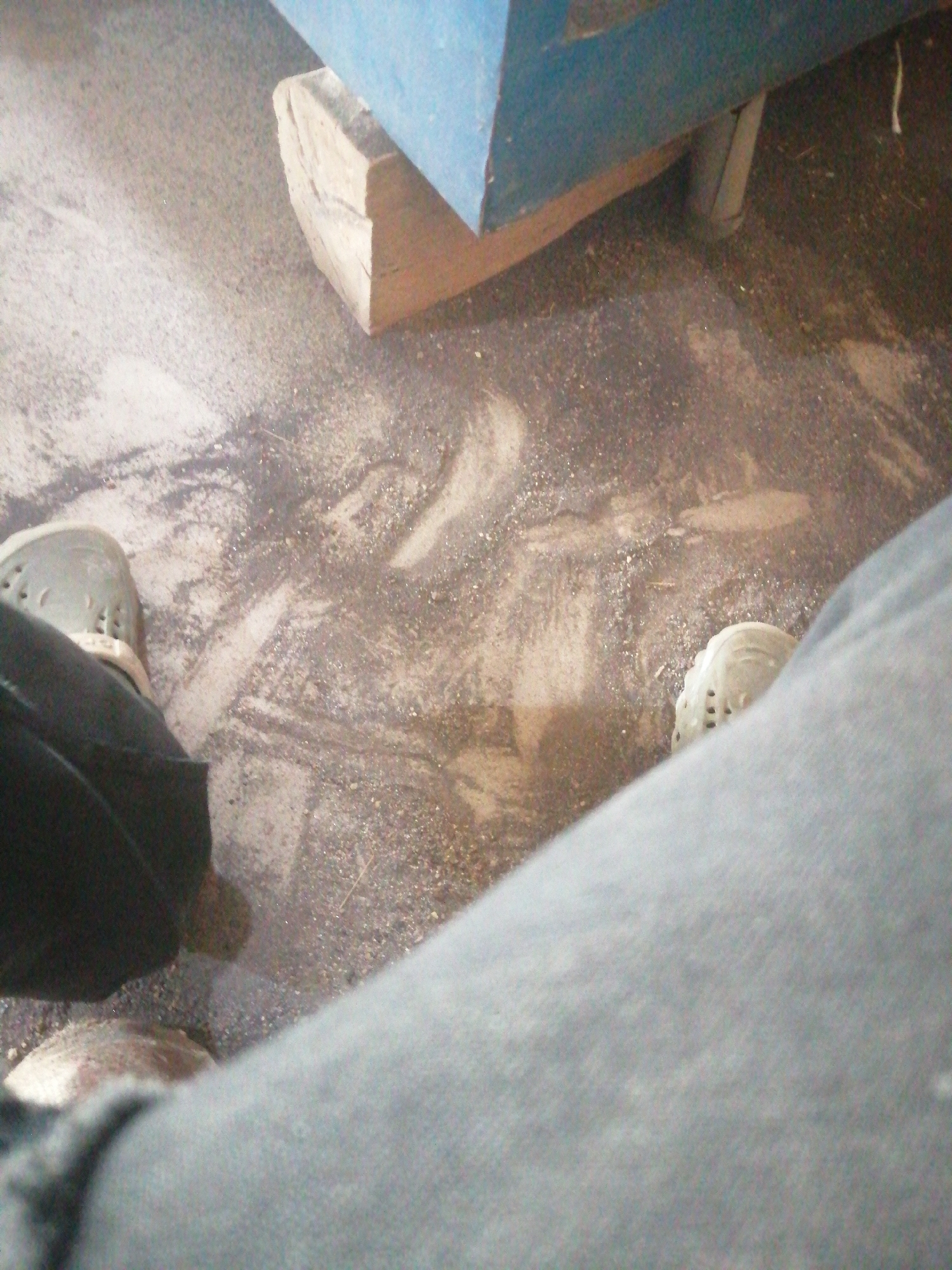 The image depicts a ground-level view of a floor covered in dirt and sawdust, with two pairs of feet visible. One person is wearing dark pants and light-colored perforated shoes, with their left foot angled towards the left edge of the frame. The other person is wearing light blue jeans and white perforated shoes, with their right foot in the lower right portion of the frame. In the background, a blue-colored structure, possibly a table or bench, is supported by a wooden block. The scene appears to be an indoor workshop or construction area. The image does not provide any specific indicators to place it within the city of Jos, Nigeria.