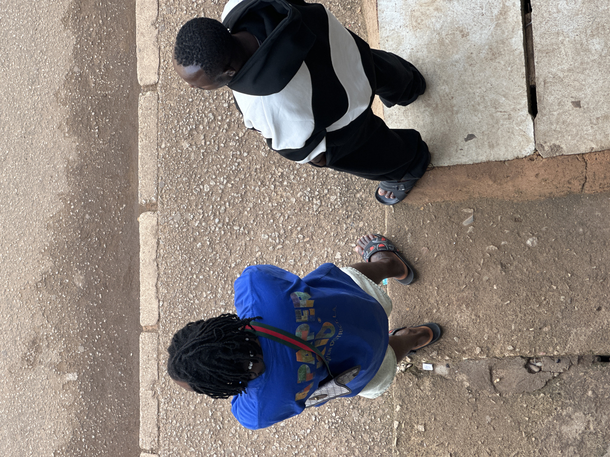 The image shows an overhead view of two young Black men standing on a dirt and concrete pathway. The man on the left is wearing a blue t-shirt with the words "PAPER" and "OWER DISTRICT, L.A." printed on the back, light-colored shorts, and black slides with red accents. He has dark, braided hair and a small crossbody bag. The man on the right is wearing a black and white horizontally striped hoodie, black pants, and black slides. They are both facing away from the camera and appear to be casually standing. The environment includes a rough, textured ground surface with scattered small rocks and debris. The image is taken in the city of Bukuru, Nigeria.
