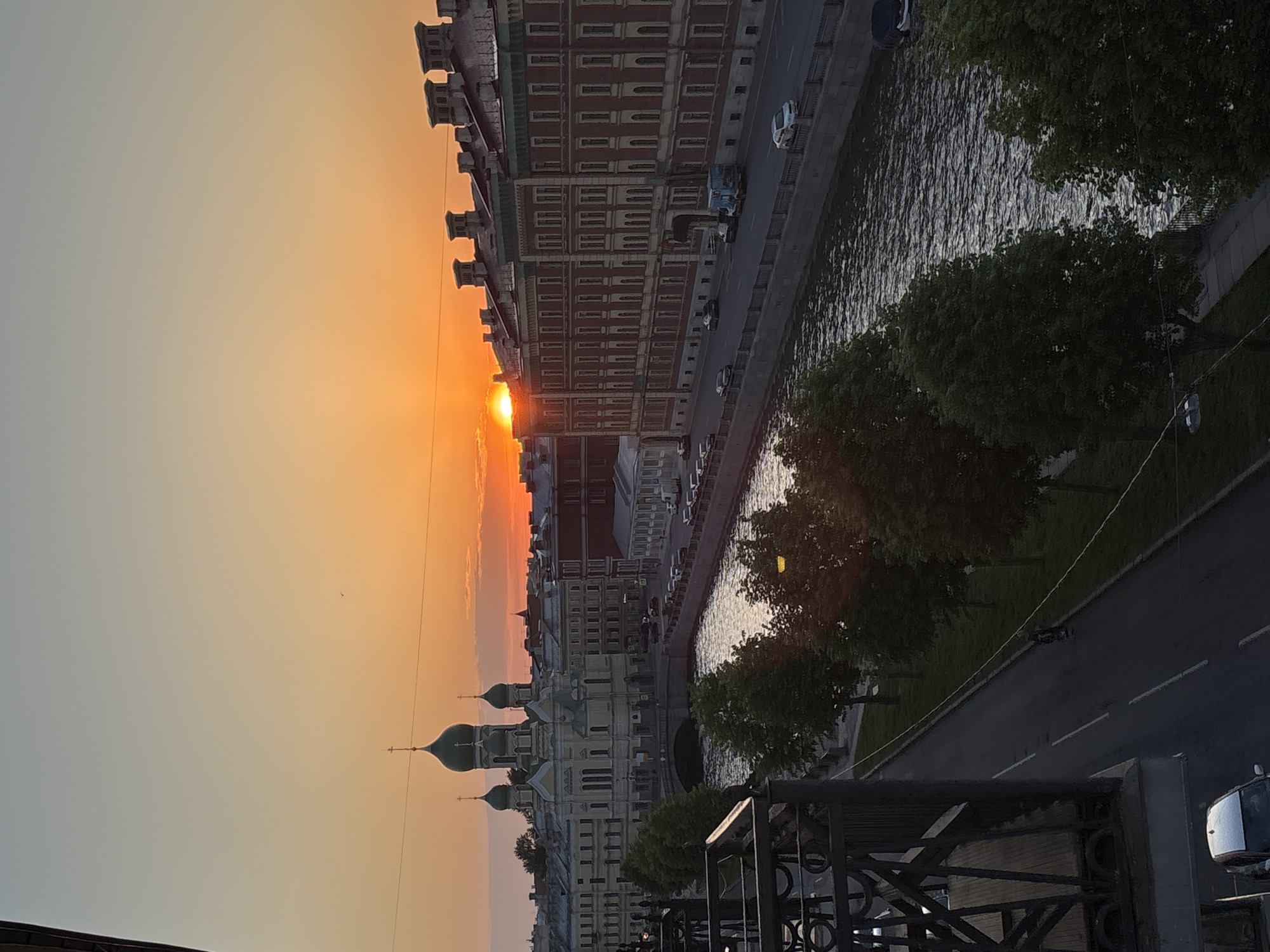 The image shows a view of Saint Petersburg, Russia, at sunset or sunrise. The sun is low in the sky, casting a warm orange glow. Buildings line a canal or river. The buildings closest to the viewer have a traditional Russian architectural style, with green domes and ornate facades. A line of trees separates the buildings from the water. A road runs along the river, with cars parked along the street. A bridge is visible over the canal. The scene conveys a peaceful and serene atmosphere. The image is likely taken from a high vantage point.