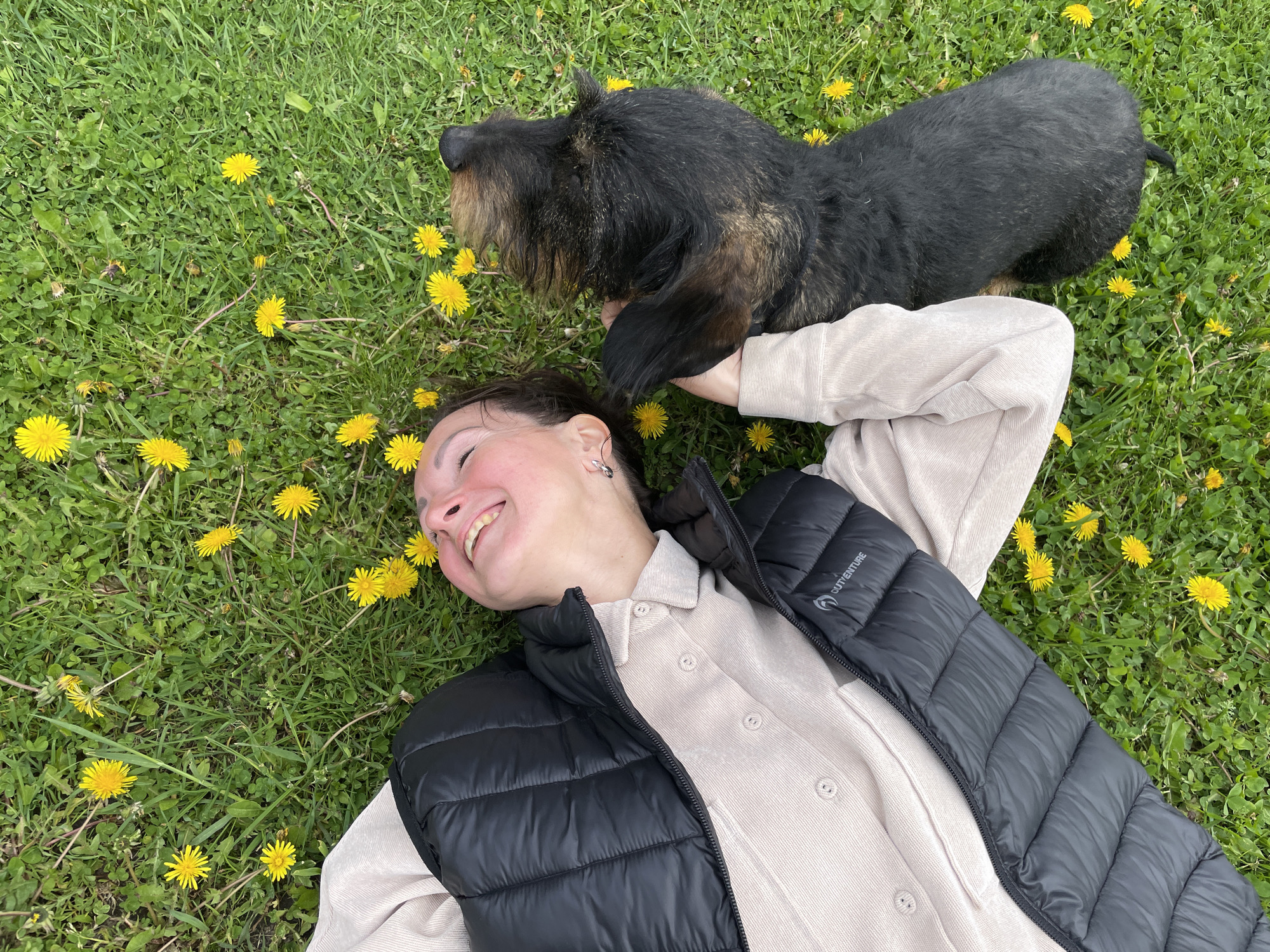 A woman is lying on a grassy field with dandelions, smiling with a dog lying on her head. The woman is wearing a light-colored button-down shirt and a black puffy vest. The dog is dark brown and black. The location is outside in a grassy field. The city of Solov'evka, Russia, is not discernible in the image.
