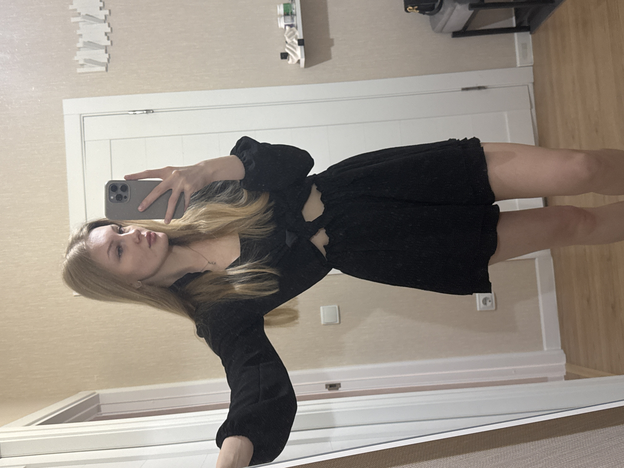 A young woman with blond hair is taking a selfie in a mirror. She is wearing a black dress with a short skirt and long sleeves. The background shows a room with light beige walls, a white door, and what appears to be a mirror frame. The room is likely in a residential setting. It is not possible to pinpoint the exact location in the city of Perm, Russia from the provided image.