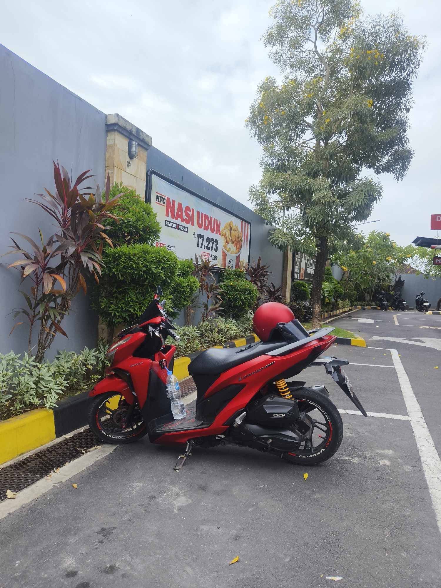 A red Honda scooter is parked on asphalt in front of a KFC restaurant with a gray wall and a large advertisement for "NASI UDUK". A red helmet rests on the scooter's rear seat. Yellow flowers are visible on a tree in the background, along with other parked scooters and a drive-thru lane. The scene appears to be a parking lot in Denpasar, Indonesia, with a overcast sky.
