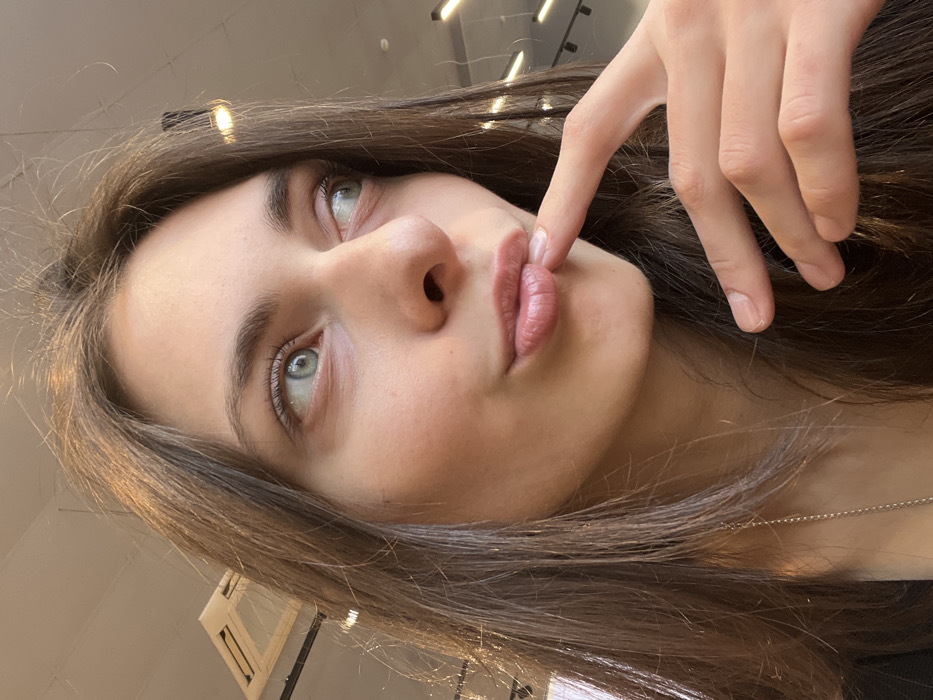 A young woman with light olive skin and brunette hair poses in a low-angle shot, her face tilted upward. Her eyes are striking with a light aqua hue. She has full lips, lightly tinted with a rosy color, and her expression suggests playful thought. A finger from her right hand gently touches the corner of her mouth. The background suggests an indoor setting with modern lighting. The overall image presents a casual and intimate feel.