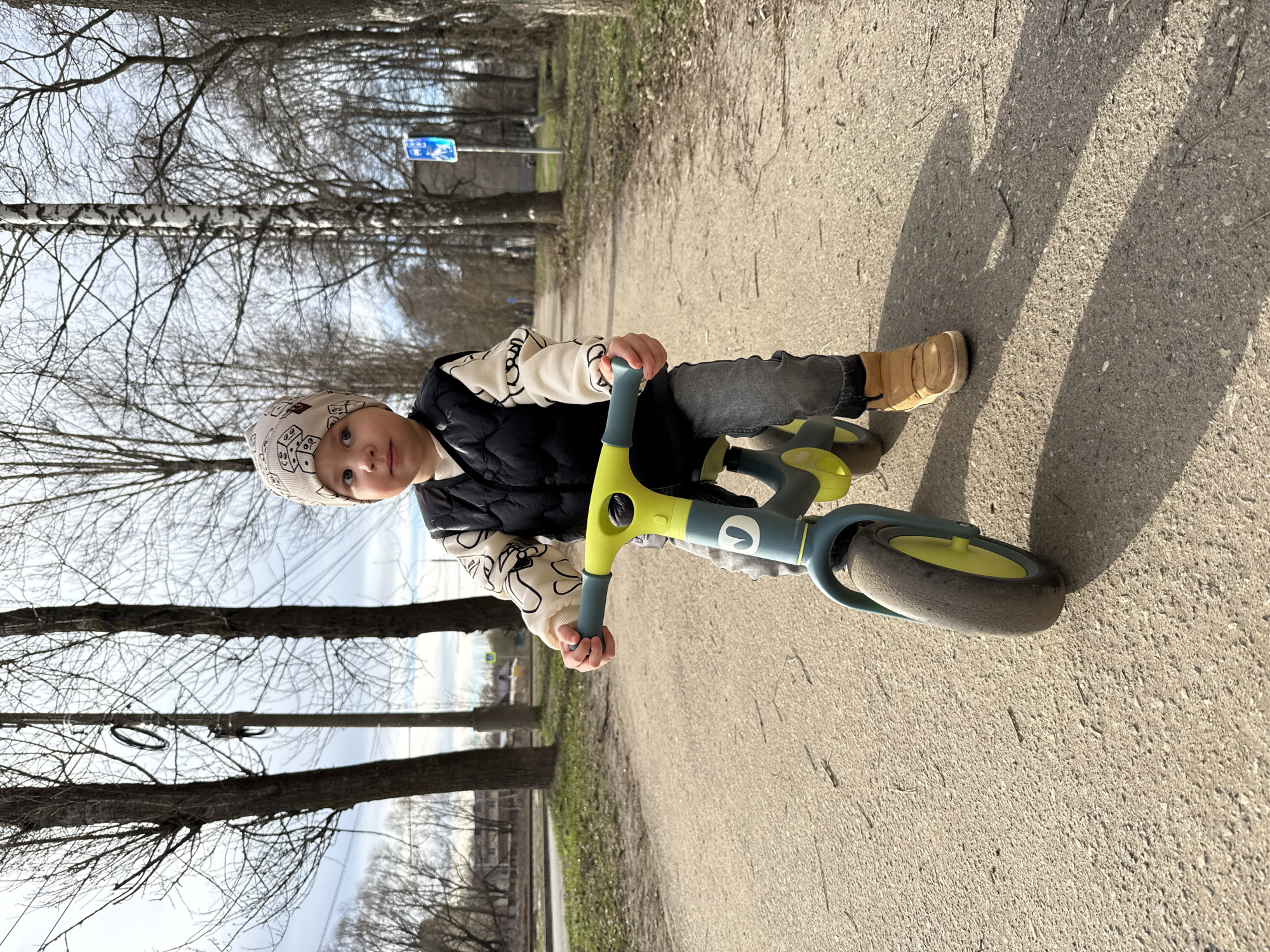 A young child is riding a balance bike on a paved path. The child is wearing a hat, a light-colored sweatshirt with a pattern, a dark vest, dark pants, and light-colored shoes. The bike is green and blue. The path is lined with trees, and in the background, there is a blue road sign. The environment appears to be a park or recreational area in Fedorino, Russia.