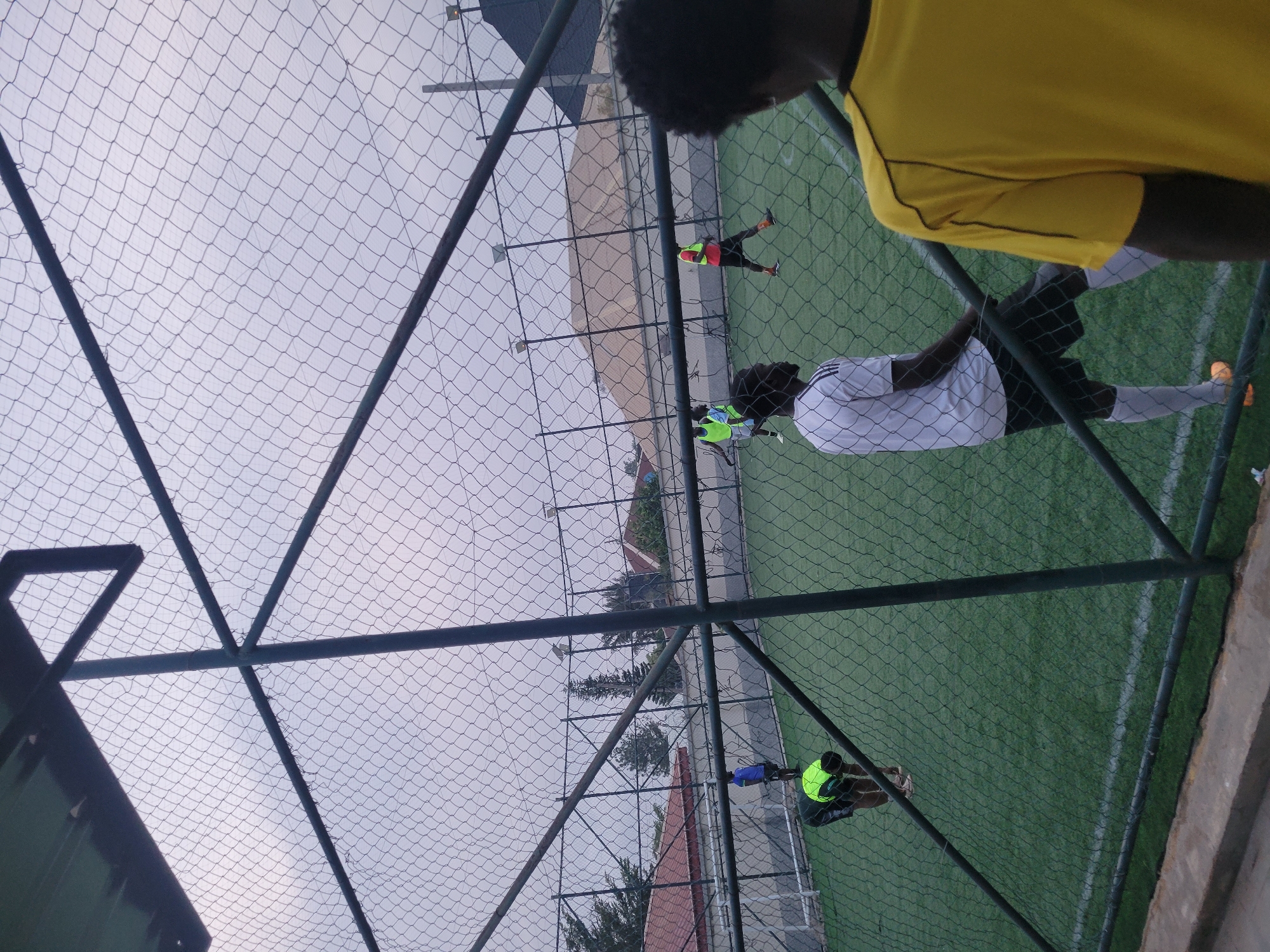 Here's a forensic description of the image:
**Scene:** An outdoor artificial turf field with a chain-link fence enclosure.
**People:** Multiple individuals are visible on the field, engaged in activity, likely soccer practice or a game. One person is wearing a bright yellow jersey is partially visible in the foreground.
**Objects:** The field is surrounded by a chain-link fence supported by metal posts. A building and trees are visible outside the fenced area.
**Environment:** The sky is overcast, indicating a cloudy day.
**Location:** Unknown, Unknown. There is no information about the city and state.