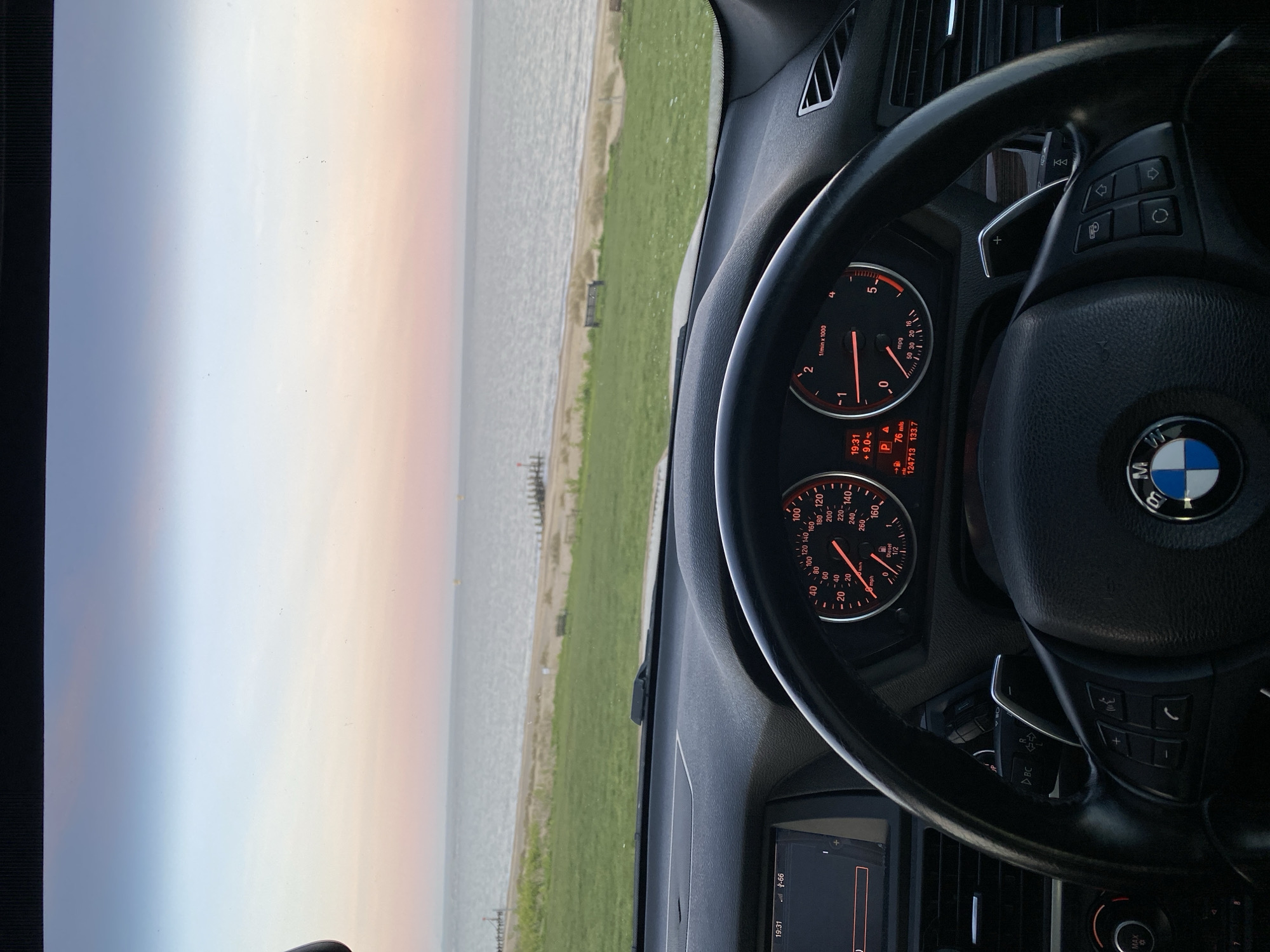 The image, taken from inside a BMW, captures a view of the sea near East Suffolk, United Kingdom. The car's dashboard and steering wheel are prominent in the foreground, with the instrument panel displaying time as 19:31, an outside temperature of 9.0°C, mileage as 76 miles, and the car's total mileage as 124713. The background reveals a coastline with a sandy beach, grass, and the sea. The sky appears overcast with diffused light. A wooden structure can be seen in the water near the shoreline.