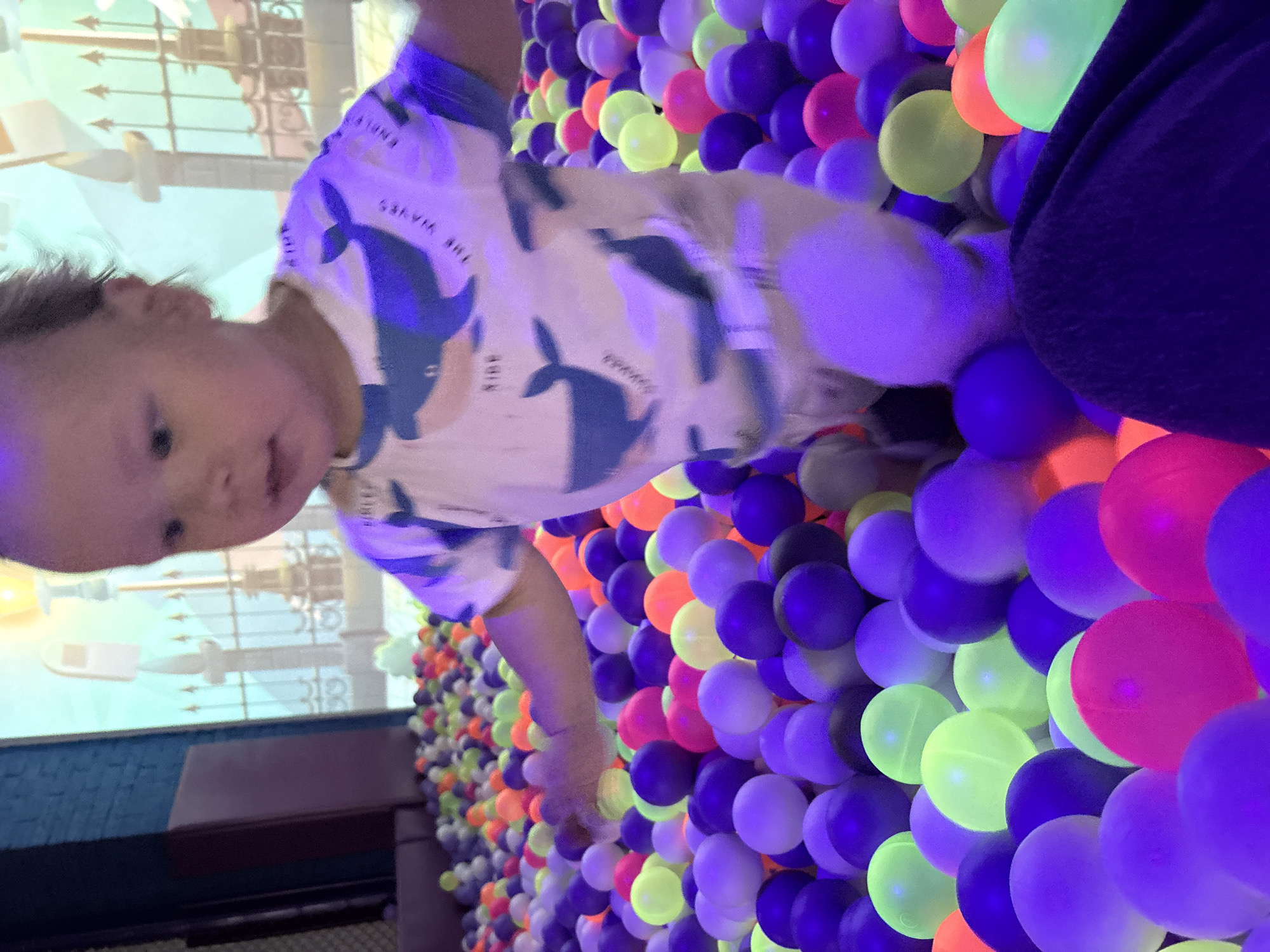 A young child is in a ball pit with multicolored plastic balls. A screen displays a cartoonish animation. The child is wearing a light-colored shirt with whale designs. The scene appears to be indoors. The location may be in Izhevsk, Russia.