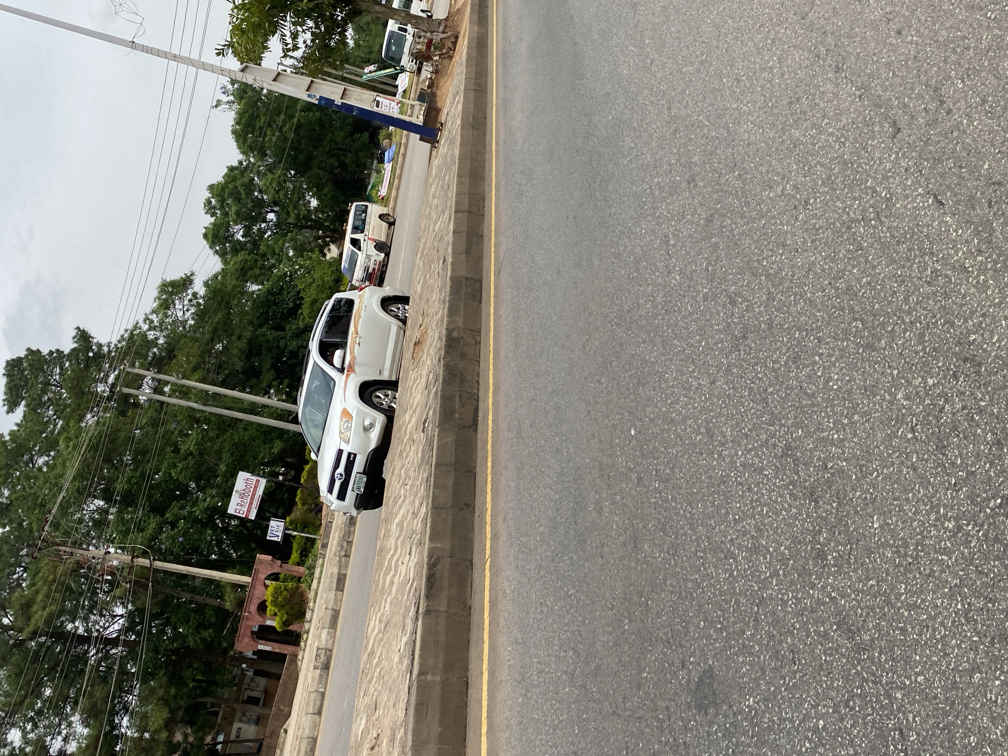An outdoor, medium-shot, eye-level, vertical, slightly angled, over-the-shoulder shot captures a cloudy, overcast day on a paved road in Jos, Nigeria. The foreground is dominated by the rough texture of the asphalt road, with a distinct yellow line running along the left edge. To the right, a concrete curb separates the road from a sidewalk paved with rectangular interlocking blocks.
Beyond the sidewalk, several vehicles are visible. A white Toyota RAV4 SUV is parked on the sidewalk, facing left, with its front wheels slightly turned. A white van with a red and white stripe is parked behind it, also facing left. Further down the street, another white van is visible.
In the background, a row of lush green trees lines the scene, with some interspersed buildings. A prominent brick archway is visible on the left, adorned with greenery. A tall utility pole with numerous wires stands on the right side of the frame, alongside a blue and white signpost. The overall impression is one of a typical urban street scene in Jos.