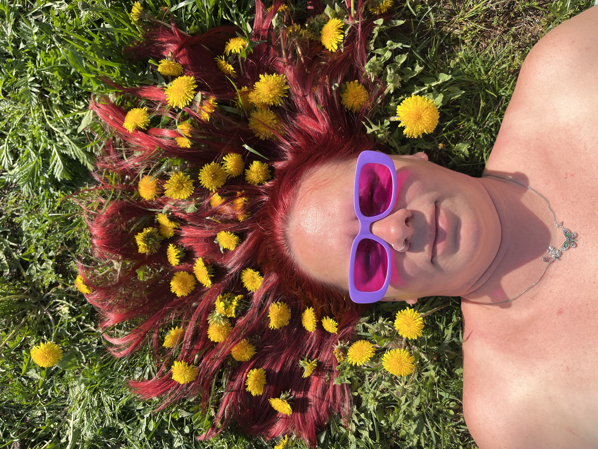 A top-down, eye-level shot shows a person lying on green grass with dandelions interspersed in the grass and arranged within the person's red hair. The person wears purple-framed sunglasses with pink lenses. The person's skin is fair and appears to be slightly sun-exposed. A thin, silver necklace with a butterfly pendant is visible on the person's neck. A silver nose ring is also visible. The green grass indicates an outdoor, grassy area. No buildings or typical markers of a city are visible in the image, so identifying Chaikovsky, Russia as the location is impossible based on this single image.