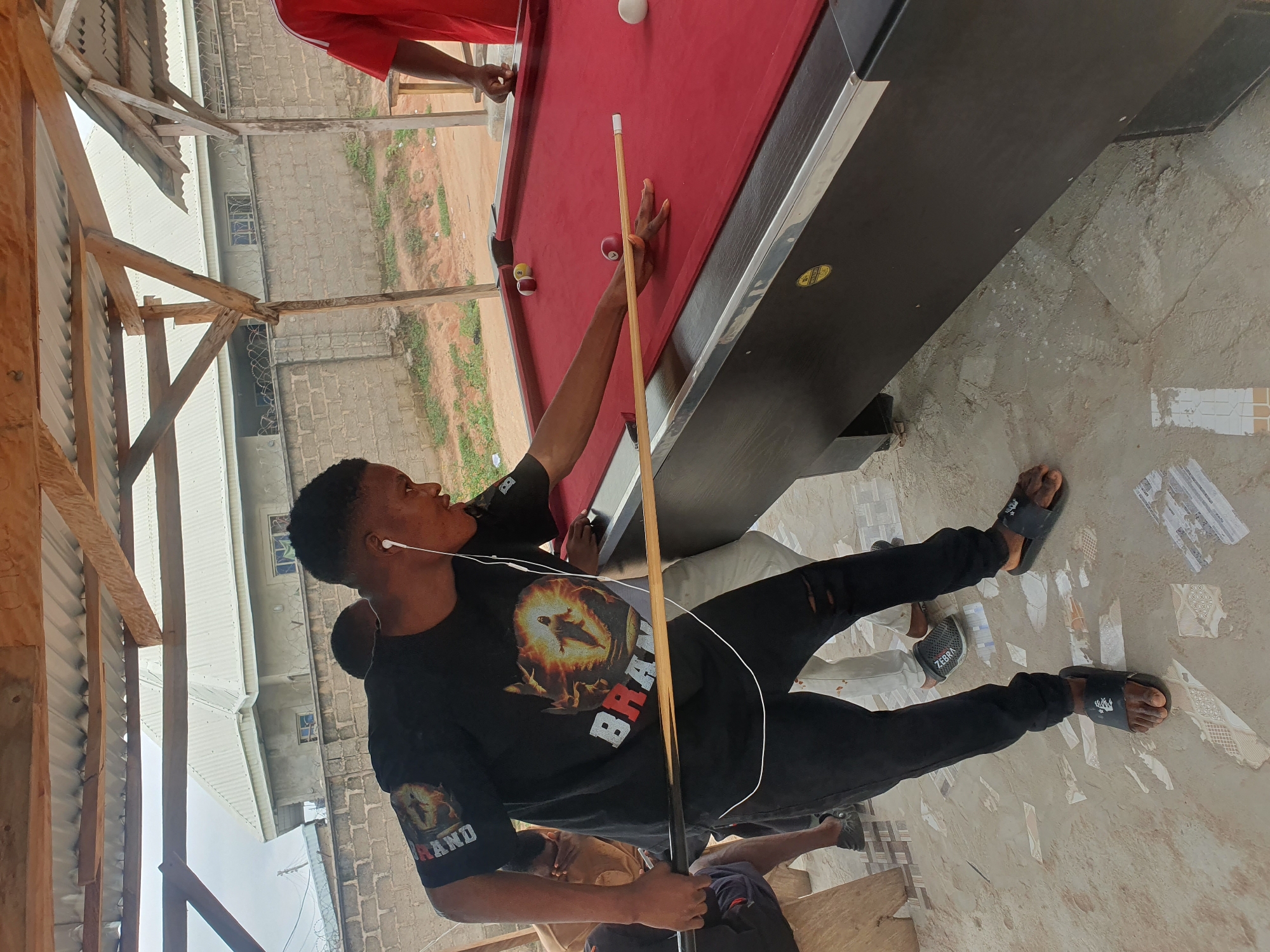 The image depicts an outdoor scene in Lokoja, Nigeria, under a corrugated metal roof supported by wooden beams. The foreground features a red billiard table with several balls scattered across its surface. A young man wearing a black t-shirt with a graphic print and ripped black pants is actively playing pool, holding a cue stick. He has earphones in his ears and is focused on the game. Other individuals are partially visible in the background. The ground is a mixture of sand and broken tiles, characteristic of an informal outdoor setting. Beyond the immediate area, a brick wall and buildings can be seen, suggesting a semi-urban or residential environment.