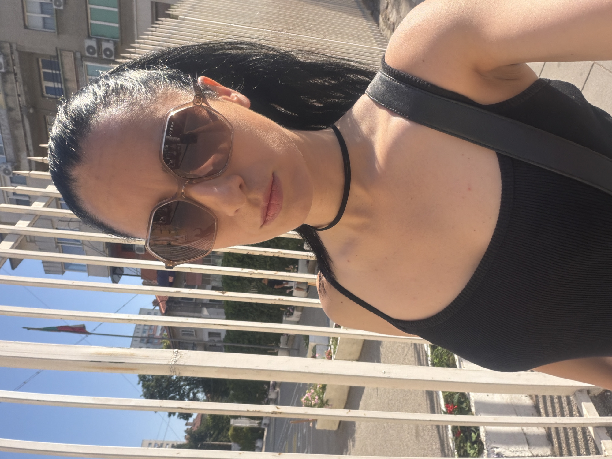 The image depicts a woman standing outdoors, possibly in the city of Pleven, Bulgaria, based on the presence of a Bulgarian flag visible behind a white metal fence. The woman, with dark hair pulled back into a ponytail and wearing large, tan-colored sunglasses and a black tank top, is looking towards the camera. A thin black choker necklace is visible around her neck. She is partially obscured by the fence, which runs vertically through the frame. In the background, buildings and trees are visible under a clear blue sky. The scene appears to be a public space, with a street and planters with flowers also present. The lighting suggests it is daytime.