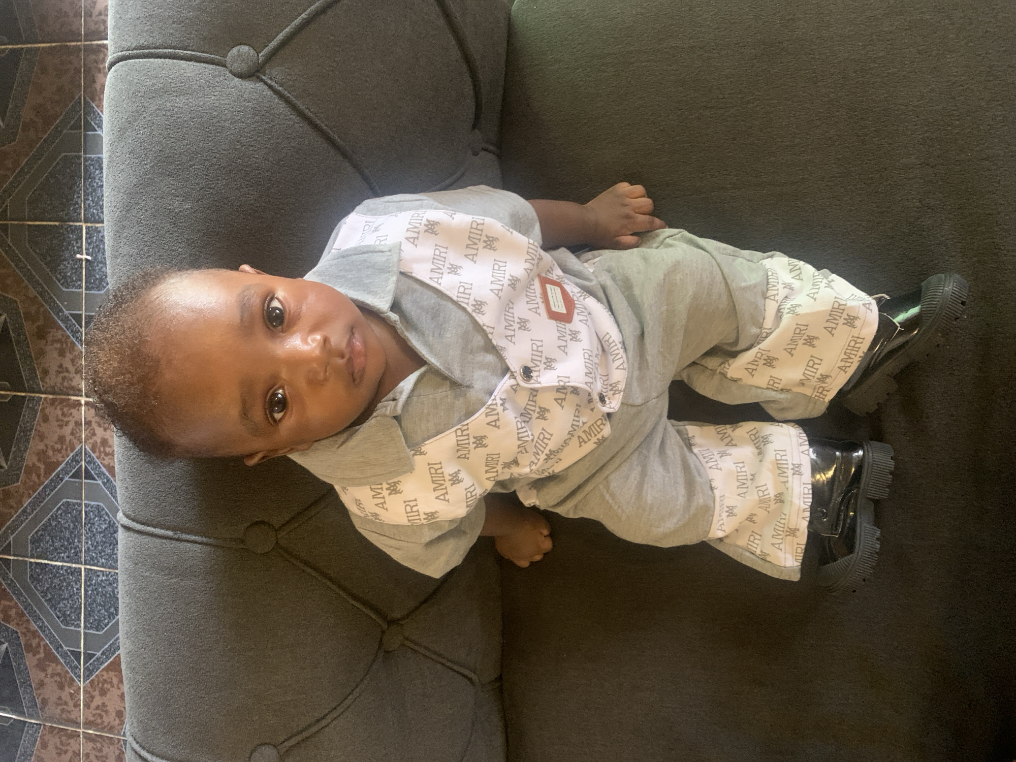 The image captures a baby or toddler lying on a dark grey upholstered sofa with button tufting. The child is looking directly at the camera with a neutral, observant expression and wide, dark eyes. They are dressed in a light grey long-sleeved top with a collar and matching pants. White sections of the outfit, including the shirt's front panel, collar, and side stripes of the pants, are patterned with repeated instances of the text "AMIRI" and a stylized "M" logo. The baby is also wearing black, chunky-soled boots. To the left of the sofa, a tiled floor with brown, black, and grey geometric patterns is visible, indicating an indoor setting. The scene appears to be lit by natural light during the daytime. The photo is rotated 90 degrees clockwise. The location is Barakin Akawo, Nigeria.