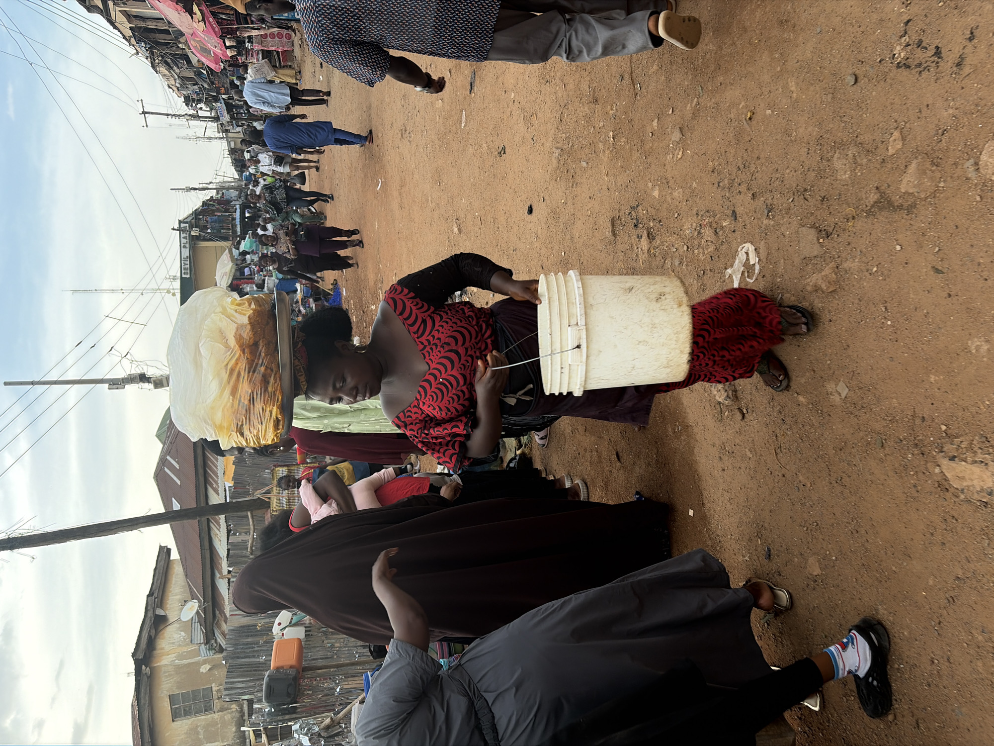 This image captures a bustling street market scene in Jos, Nigeria, characterized by a lively atmosphere and unpaved, reddish-brown ground. The time of day appears to be daylight, possibly morning or afternoon, under an overcast or partly cloudy sky, with soft, diffused lighting.
In the foreground, a woman wearing a red and black patterned off-shoulder top and a red skirt is prominent. She carries a large, light-colored plastic bucket in her right hand, from which a wire or string extends, possibly holding an item. On her head, she expertly balances a large quantity of goods, likely food items, encased in a translucent white plastic bag, indicating her role as a vendor or transporter of goods. Her expression is focused, demonstrating the physical labor involved in her activity. She wears dark sandals.
Around her, numerous other individuals populate the scene, suggesting a busy commercial area. People are seen walking, standing, and interacting, though specific activities are not clearly discernible for most. In the mid-ground, a man in a blue patterned shirt and grey trousers walks away. In the lower foreground, parts of other individuals are visible, including a person in a dark garment with one foot clad in a black clog with white socks.
The setting features buildings on both sides of the street, some with corrugated metal roofs and weathered facades, typical of urban areas in the region. Utility poles with multiple electrical wires crisscross above the street, adding to the urban landscape. A notable detail is the sign "SEVIL PLAZA" visible on a building in the mid-ground, hinting at commercial establishments. The ground is uneven, scattered with small stones and pieces of debris, reflecting the well-trodden nature of the path. The overall impression is one of everyday commerce and community life in an active market environment.