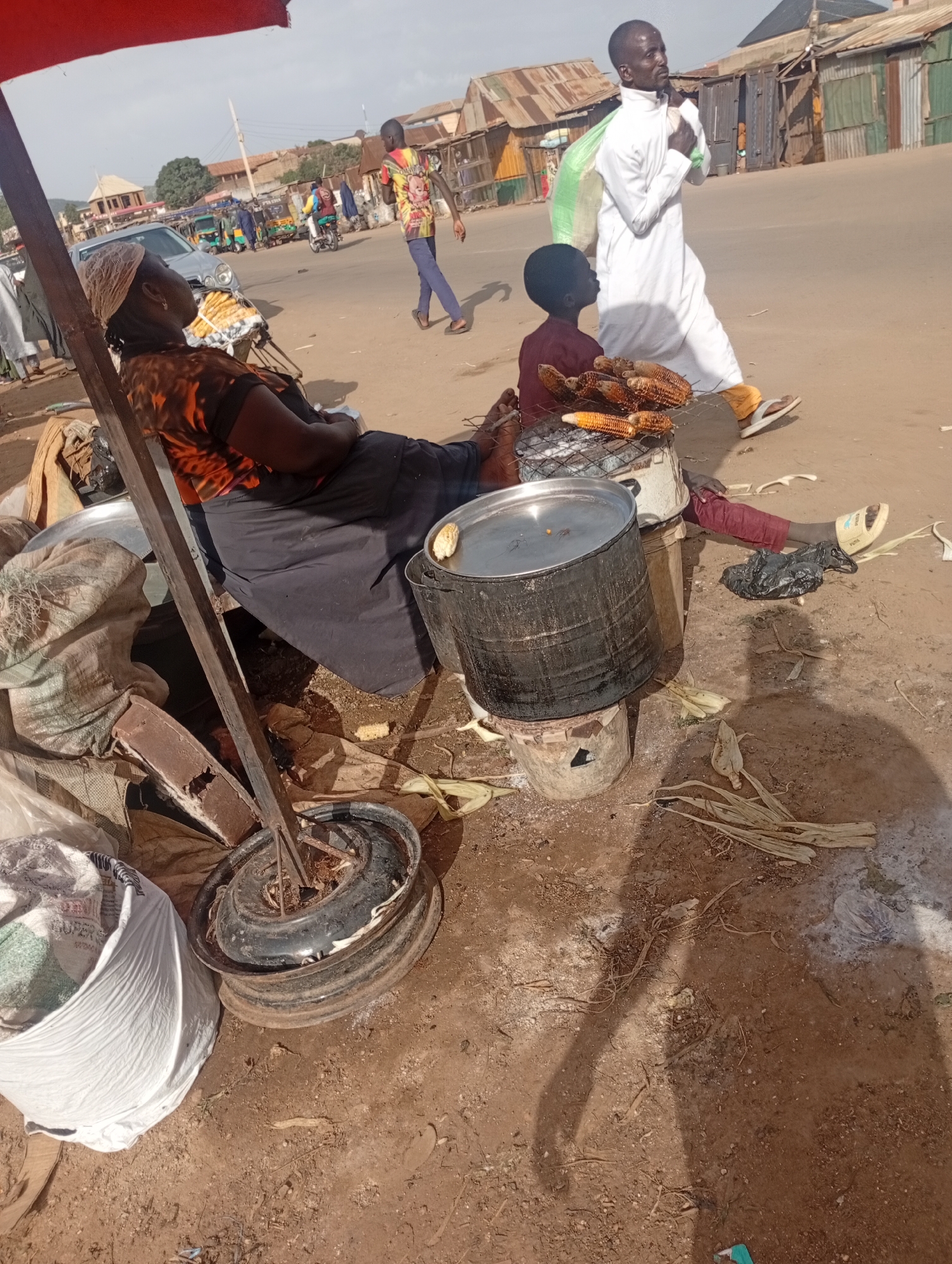 In Rafin Par, Nigeria, a woman sits under a red umbrella, her back to the camera, wearing an orange and black patterned shirt and a dark skirt. She is positioned next to a large black pot sitting on a makeshift stove, with a pile of corn on a grill to her right. A young boy in a maroon shirt sits on the ground, facing the corn. A man in a white robe walks away from the scene, carrying a large green bag over his shoulder. The ground is dirt, with scattered corn husks and debris. In the background, there are several buildings with corrugated metal roofs and a street with passing vehicles, including a car and several tricycles. The scene appears to be a street market or roadside food stall.