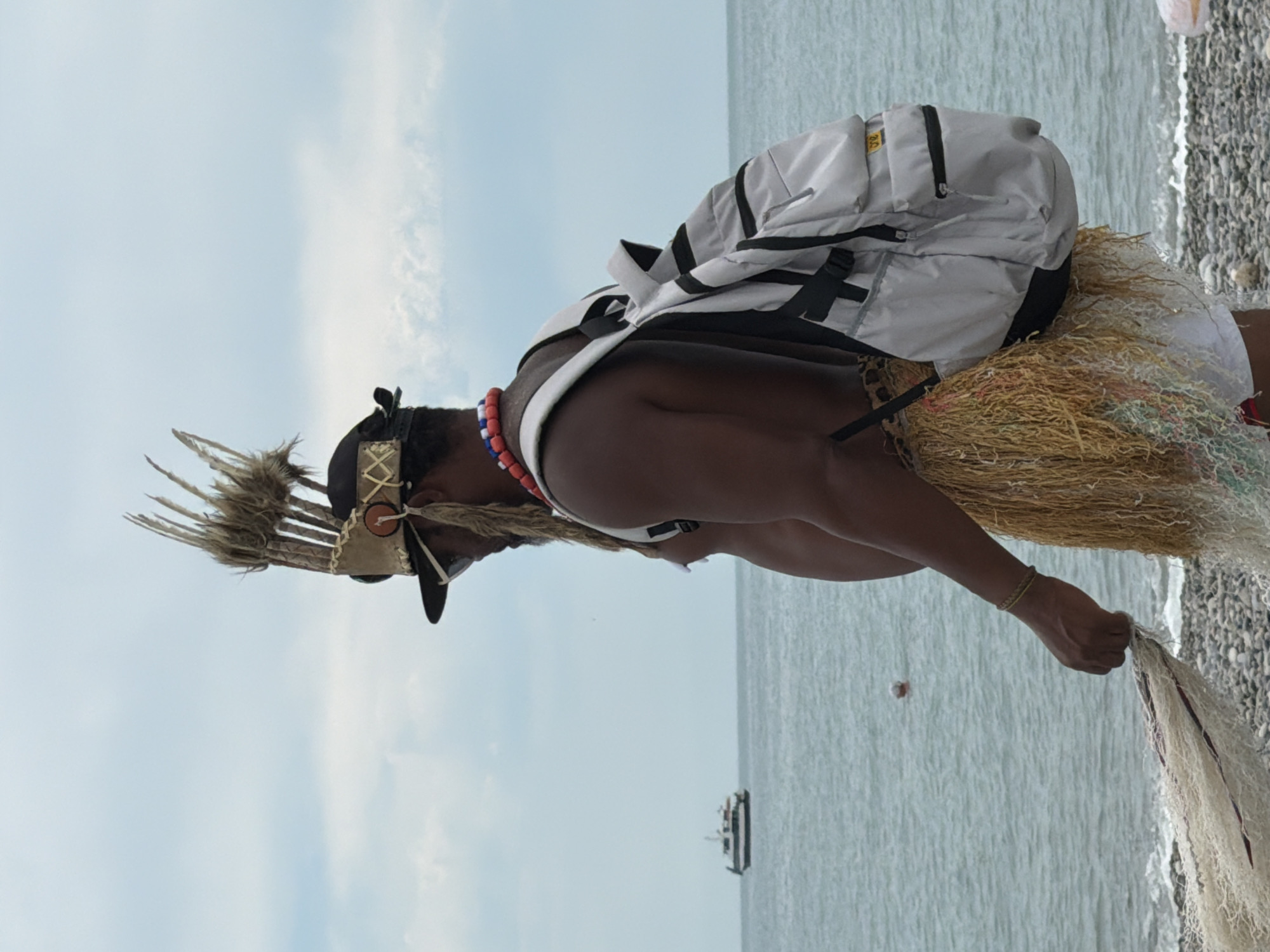 A man wearing a traditional headdress and a grass skirt stands on a pebble beach, holding a fishing net. He is also wearing a backpack. In the background, the sea stretches to the horizon, with a boat visible in the distance. The sky is overcast with clouds. The location is likely the coast of Sochi, Russia, given the presence of a pebble beach and the type of boat, which is consistent with those found in the Black Sea region.