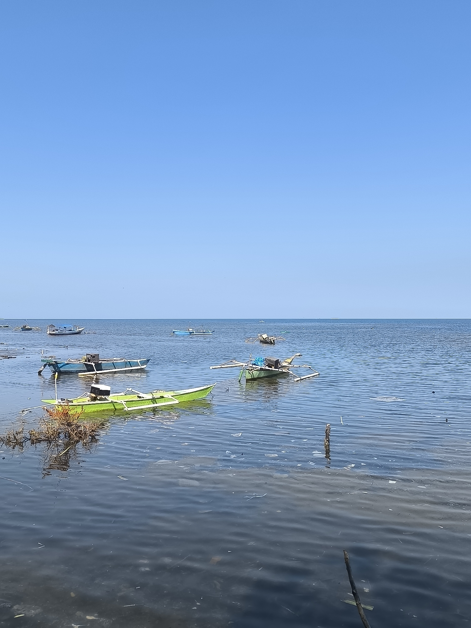 The image shows a serene body of water, likely the sea or a large lake, under a clear blue sky. Several small boats are scattered across the water. In the foreground, a bright green outrigger canoe is prominent, with some dried vegetation on its left. To its right and slightly behind it, a blue and white outrigger boat is visible. Further in the distance, more boats of various colors and sizes are present, some appearing to be anchored or moored. The water exhibits a gentle ripple, and some debris, possibly leaves or plastic, floats on the surface, particularly in the foreground. The horizon is clearly defined, separating the blue of the water from the vast expanse of the sky. This scene is consistent with a coastal or lakeside area in the city of Barru, Indonesia, likely a fishing village or a popular spot for small boat activities.