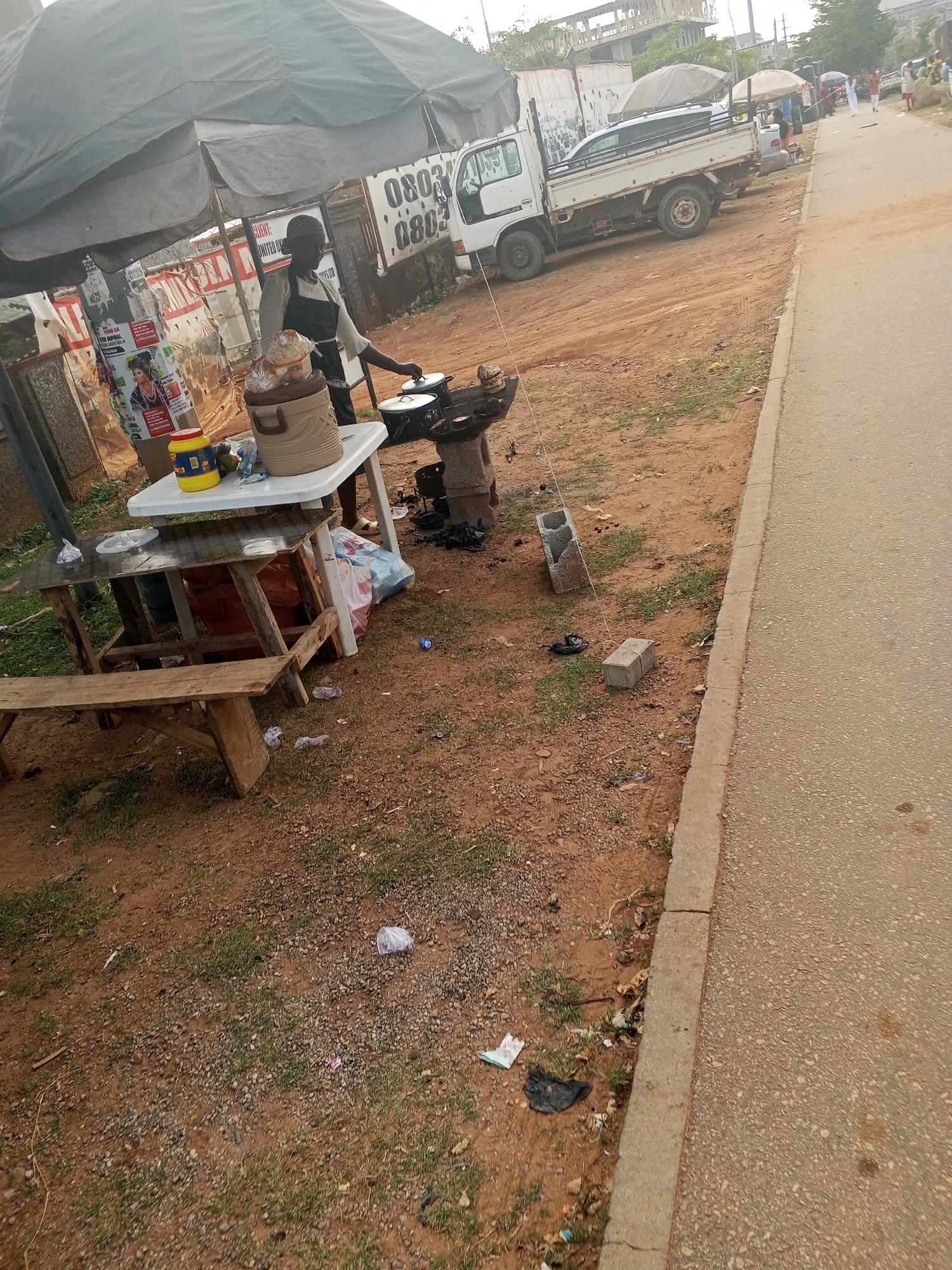 The image depicts an outdoor food vending setup in what appears to be a roadside location. A person wearing an apron is visible near a cooking stove, likely preparing food. The setup includes a table, a cooler, and cooking utensils. A covered tent-like structure provides shade. The ground is a mix of dirt, grass, and litter. A truck and other vehicles are parked nearby. The scene includes a concrete sidewalk and a paved road. Judging from the vehicle registration plates, the location is likely in Nigeria. There are no specific details available in the image to confirm that the location is in the city of Abuja.