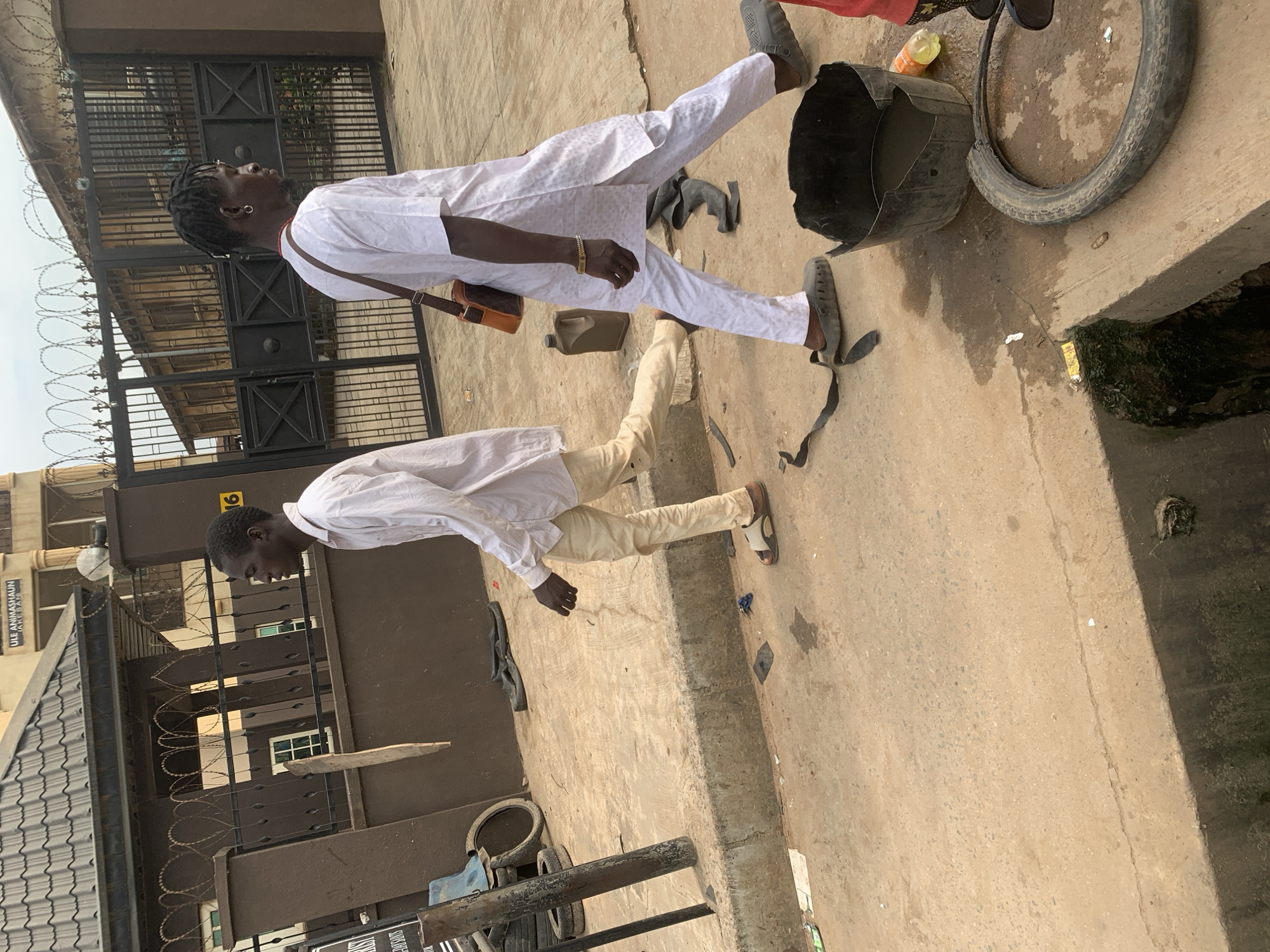 Two young men are walking side-by-side on a dirt and concrete street in Shagamu, Nigeria. The man on the left wears a light-colored long-sleeved shirt and tan pants, with open-toed sandals. The man on the right wears a white dashiki with an embroidered pattern and matching pants, also in open-toed sandals. He carries a brown messenger bag over his left shoulder. Behind them, a large black metal gate is visible, with barbed wire along the top of a brown wall. To the left of the gate, a building with a dark tiled roof can be seen. The ground is littered with debris, including a broken tire, a black bucket with water, and what appears to be a discarded plastic bottle. The overall scene depicts an outdoor urban environment with signs of street-level activity and possibly informal commerce.