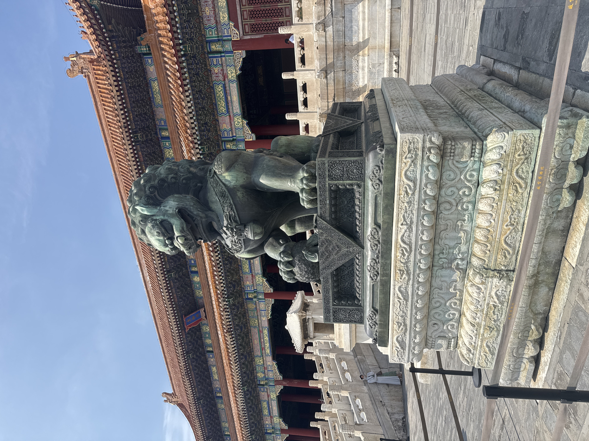 A bronze lion statue stands in the foreground, facing left, in front of a traditional Chinese palace building. The statue is dark green with intricate details on its mane and body. It rests on a large, ornate stone pedestal adorned with carvings of dragons and swirling patterns. In the middle ground, a person in a light-colored robe stands on a wide stone staircase to the left of the statue. The palace building in the background features a red tiled roof with elaborate, colorful decorations along the eaves and walls. Red pillars support the overhanging roof. The ground is paved with gray flagstones, and black stanchions with horizontal ropes are visible in the foreground, suggesting a restricted area. The sky is clear and blue. The scene is set in the Forbidden City, located in Dongcheng District, China.